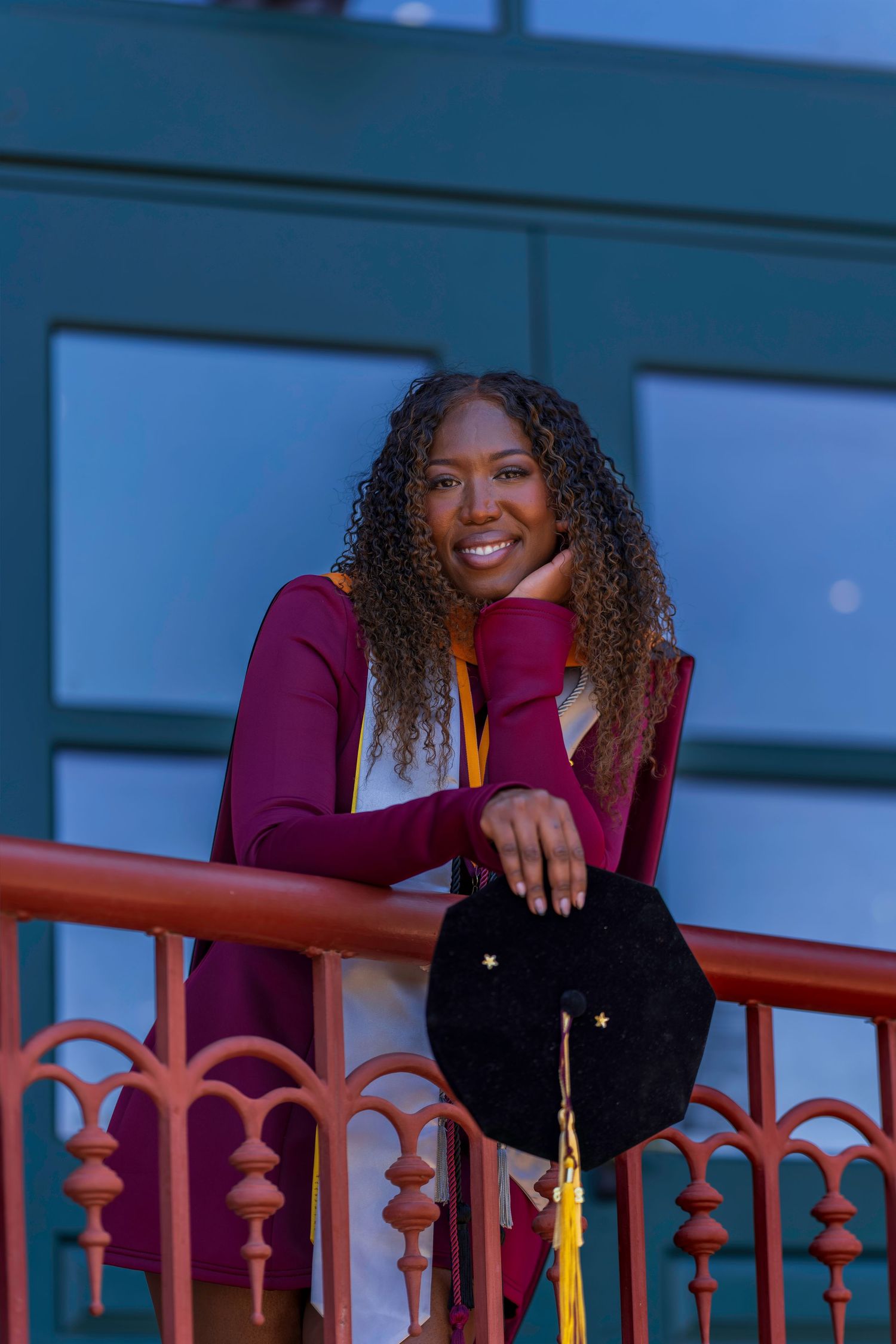 Smiling graduate in maroon gown holds cap on balcony, wearing gold honor cord at dusk.