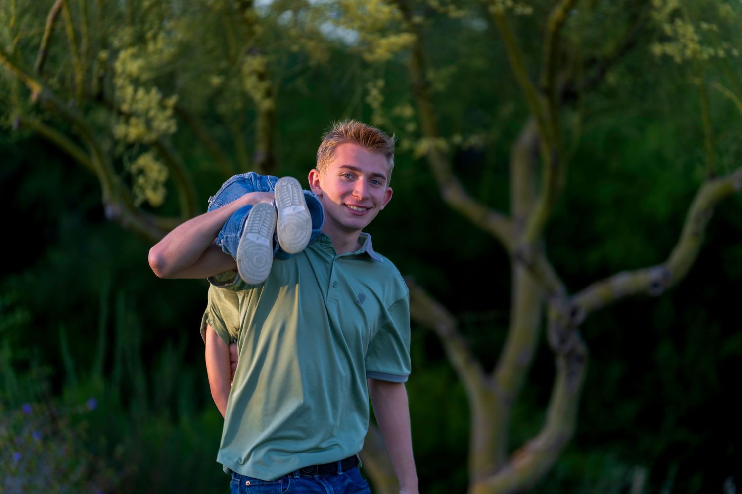 Smiling teenage boy in green polo shirt carrying someone on his back outdoors near trees.