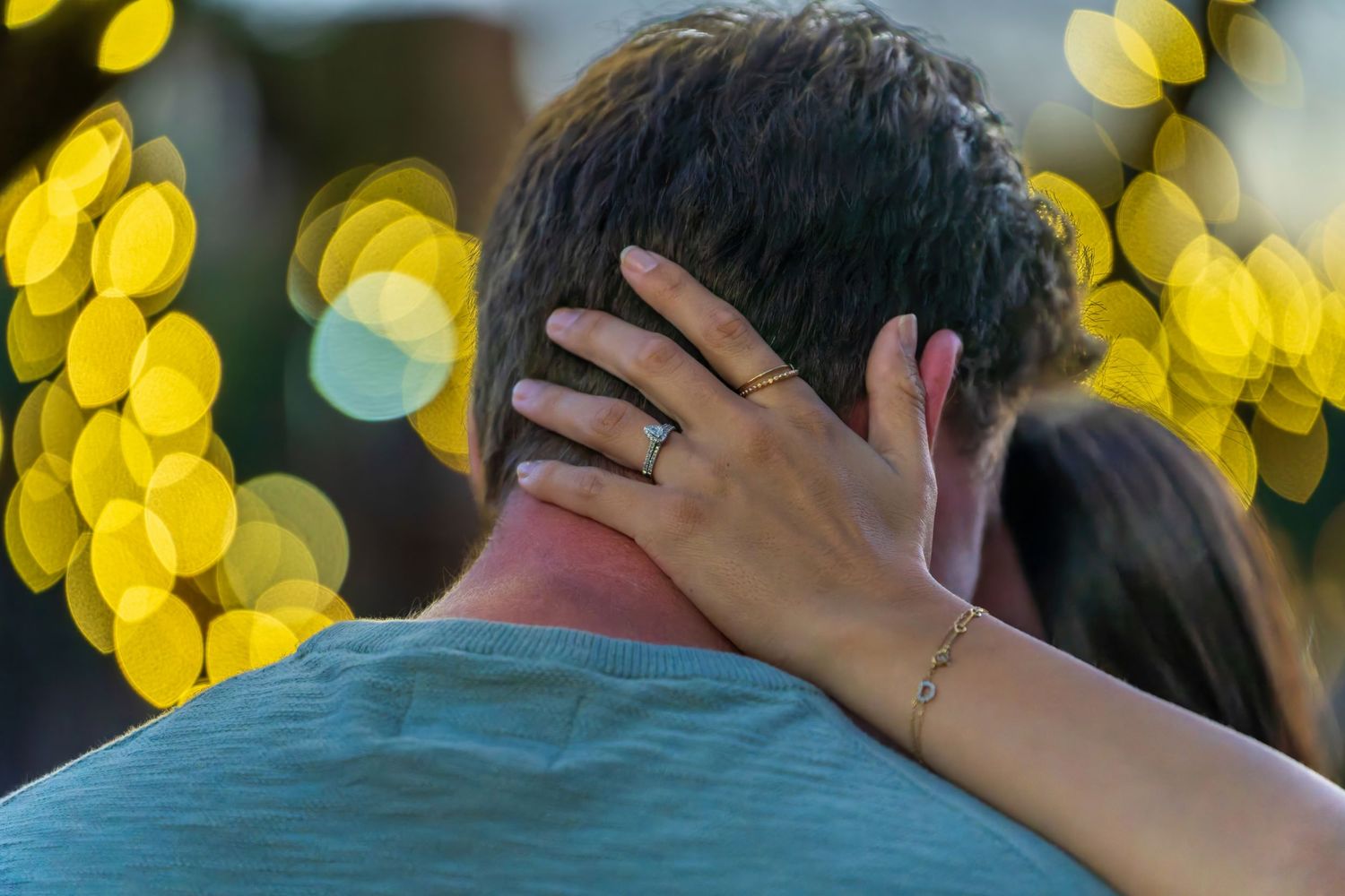 Close-up of woman's hand with engagement ring resting on man's shoulder with golden bokeh lights background.