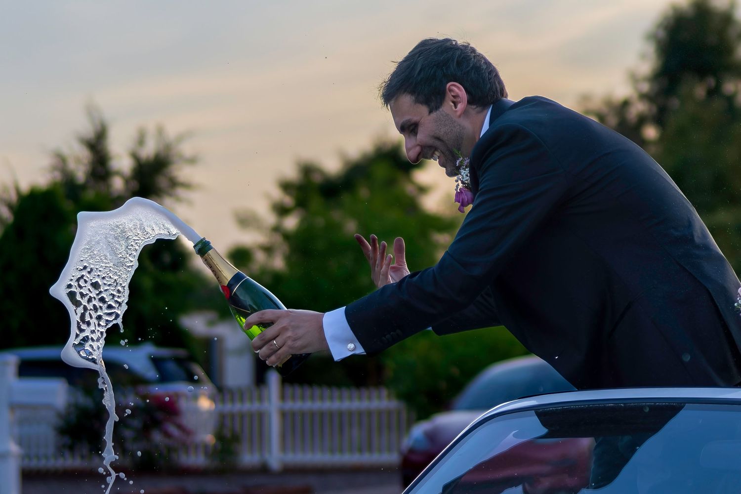 Man in suit popping champagne bottle at outdoor wedding celebration at dusk.