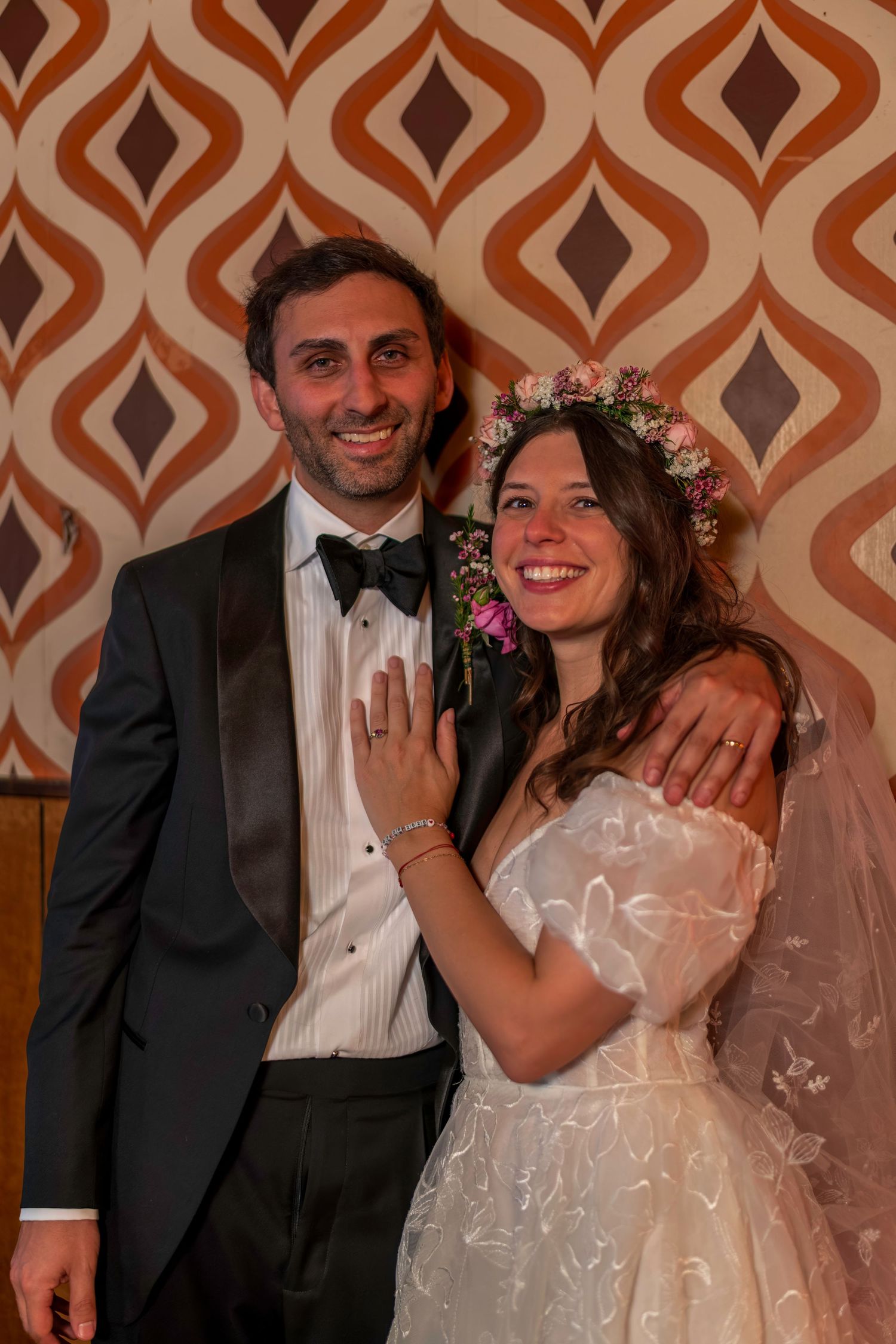 A smiling bride in a floral crown and groom in a tuxedo pose together on their wedding day.