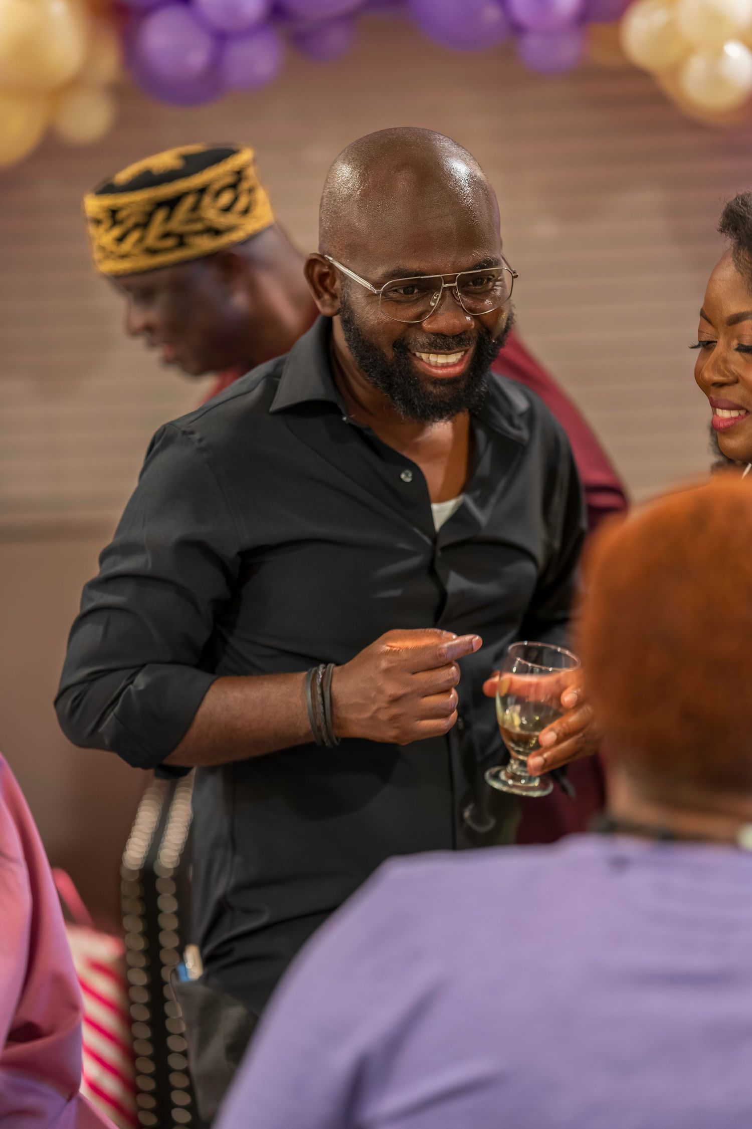 Smiling bald man with glasses holding a wine glass at a festive party with balloons in the background.