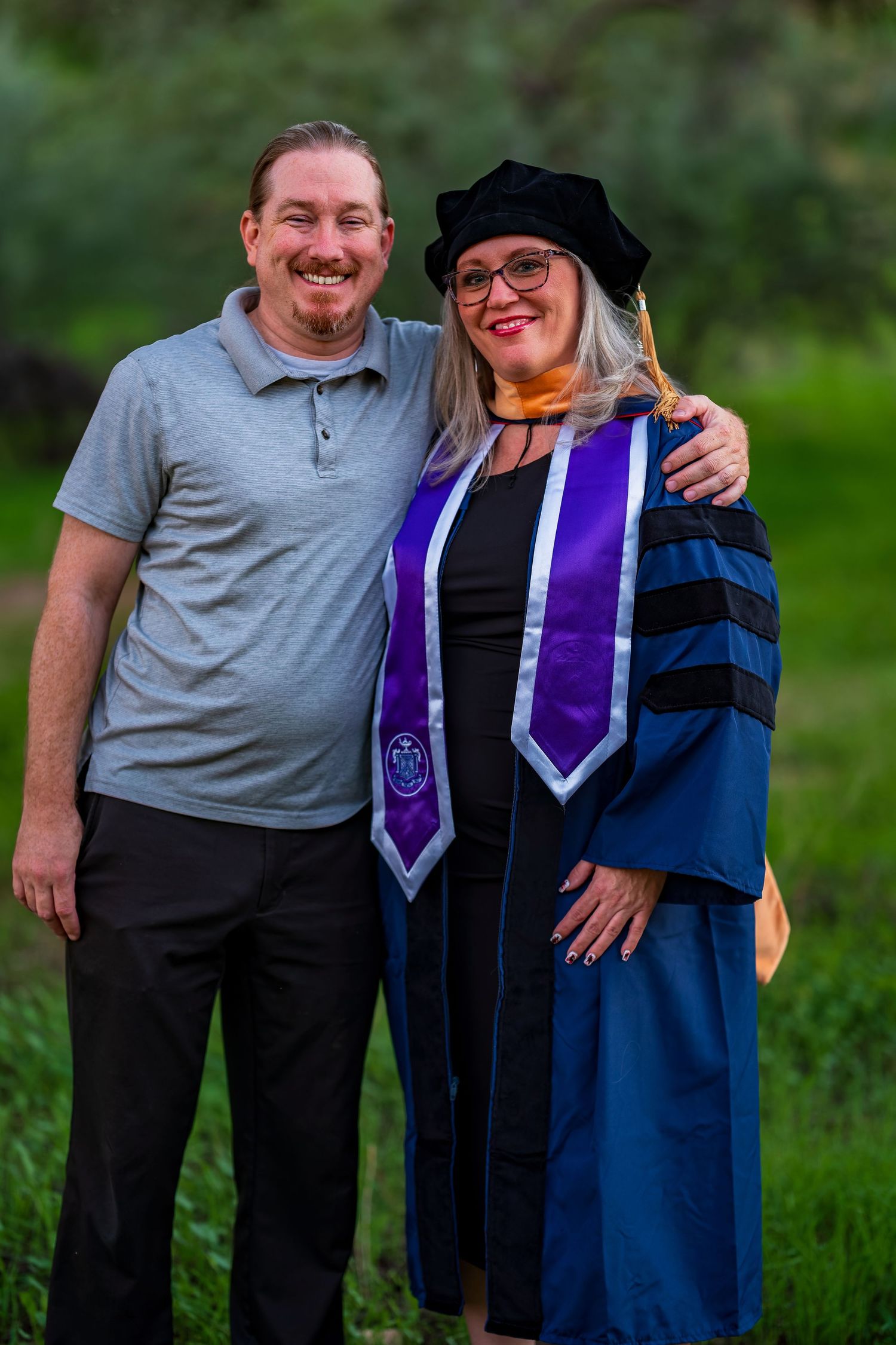 Two people celebrate graduation day together outdoors in formal attire and academic regalia.