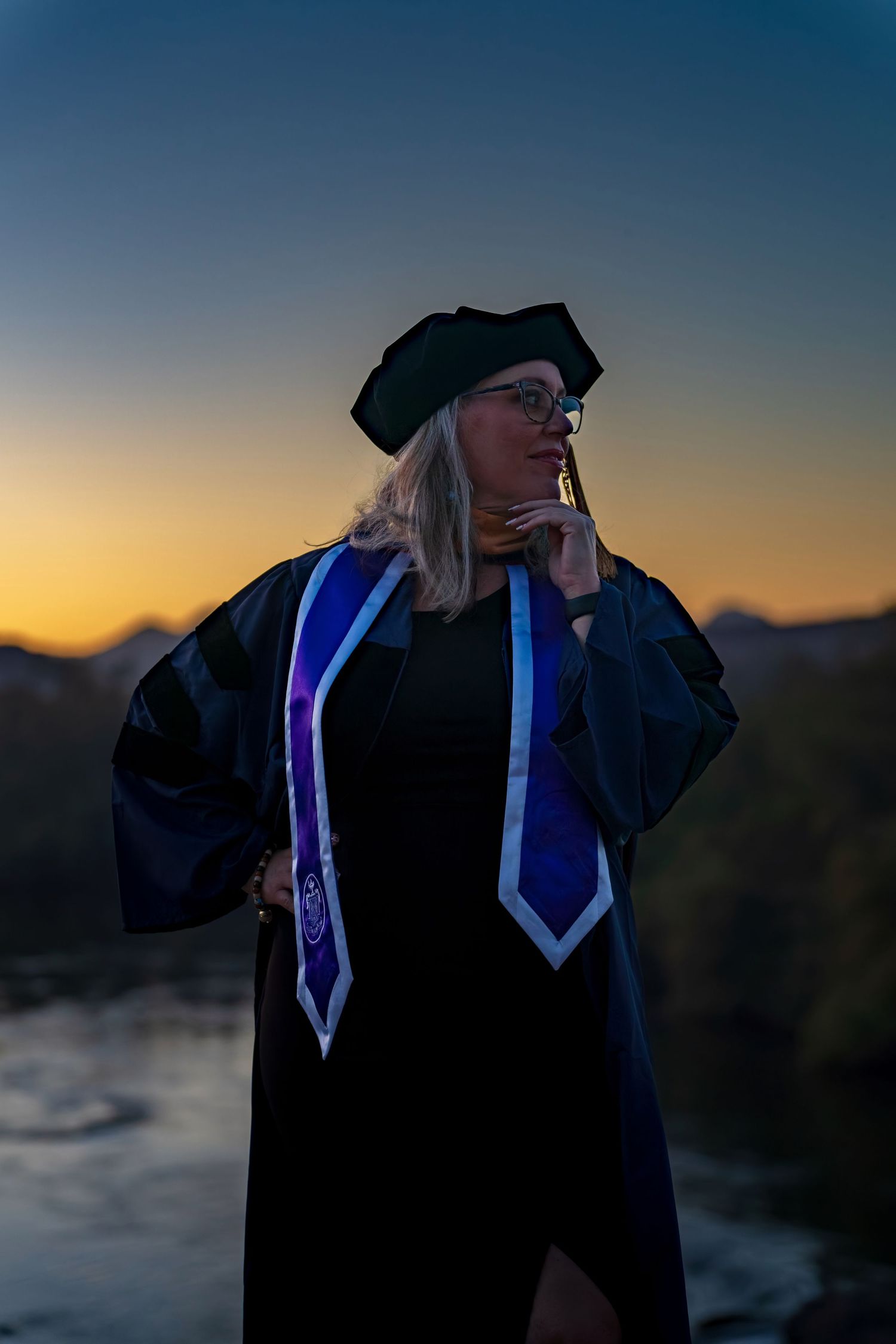 Silhouette of graduate in academic dress against dramatic sunset and water backdrop.