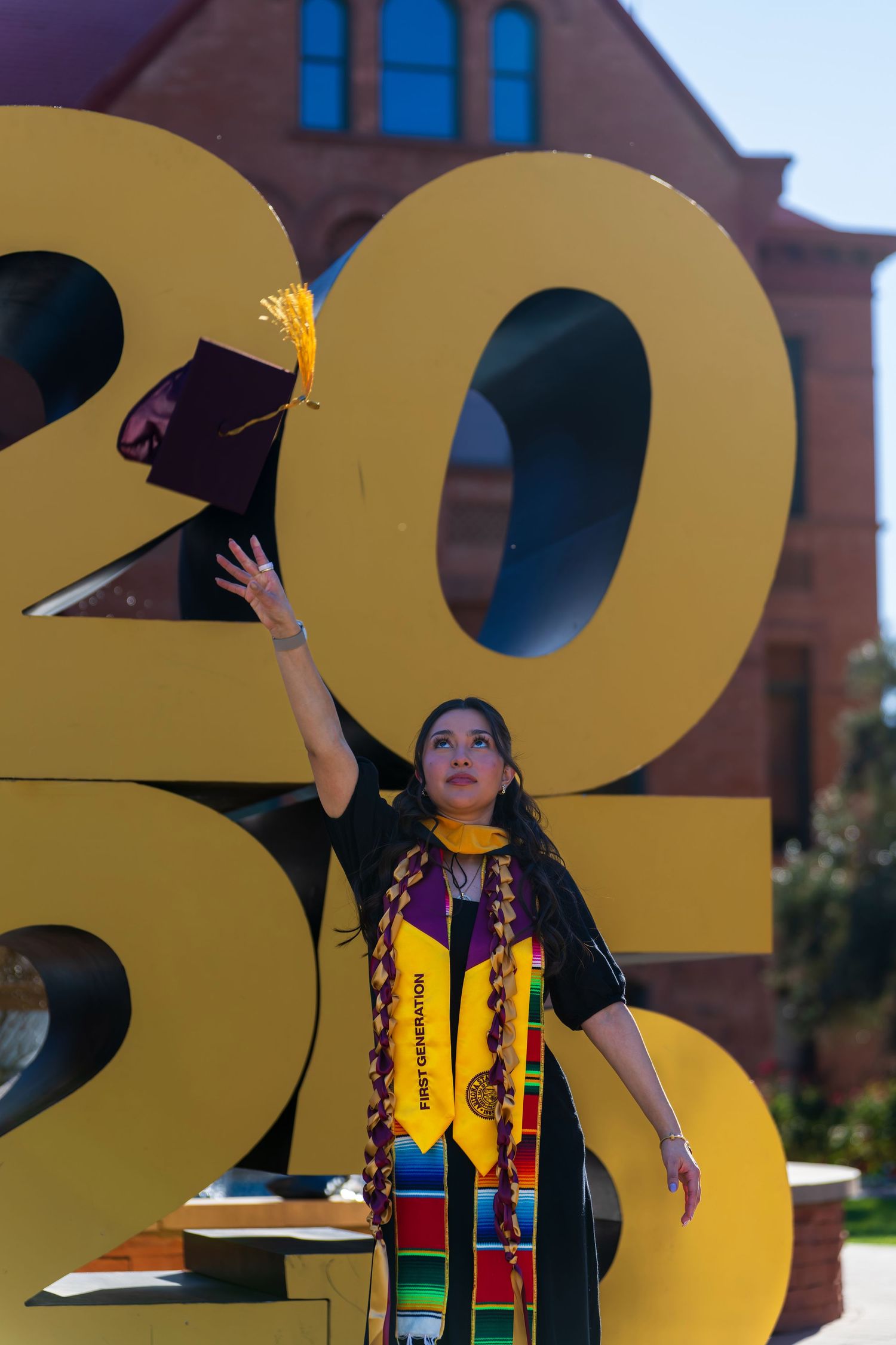 Graduate in colorful regalia poses with large gold 2023 numbers outside a brick building.