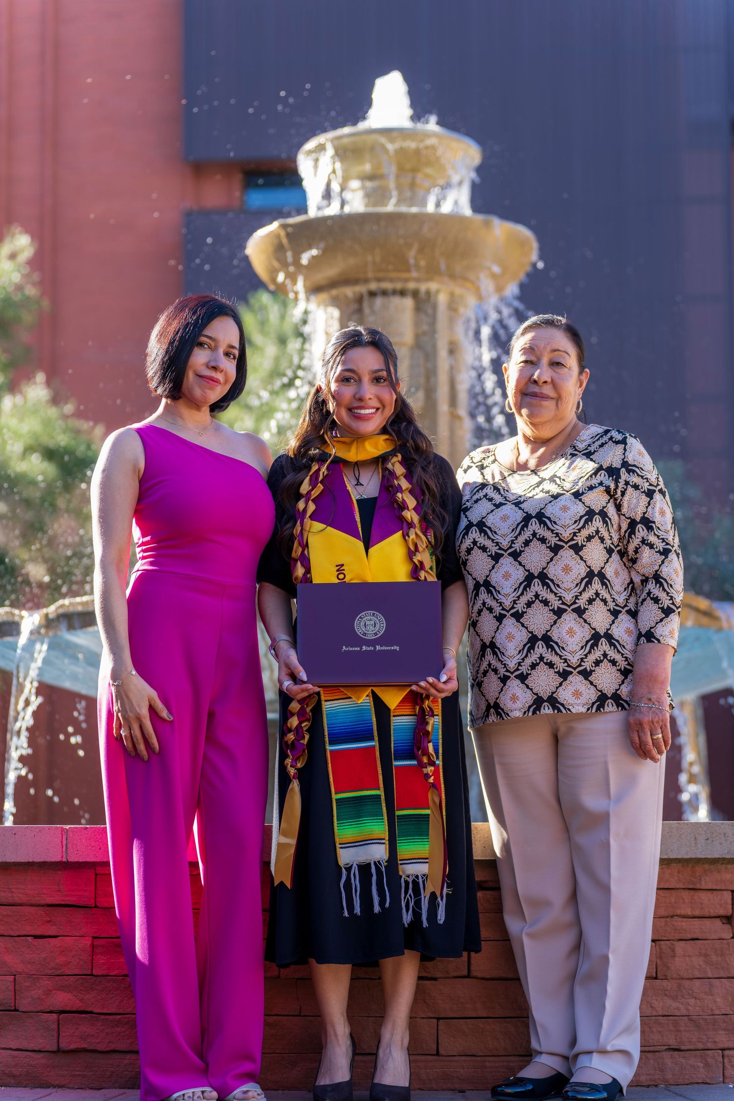 Group photo in front of a red brick building with a fountain, wearing colorful graduation attire.