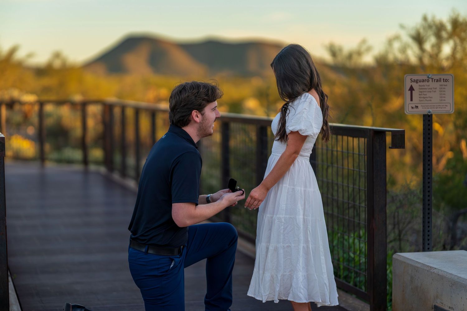 A romantic proposal scene on an outdoor bridge with desert mountains and sunset lighting.