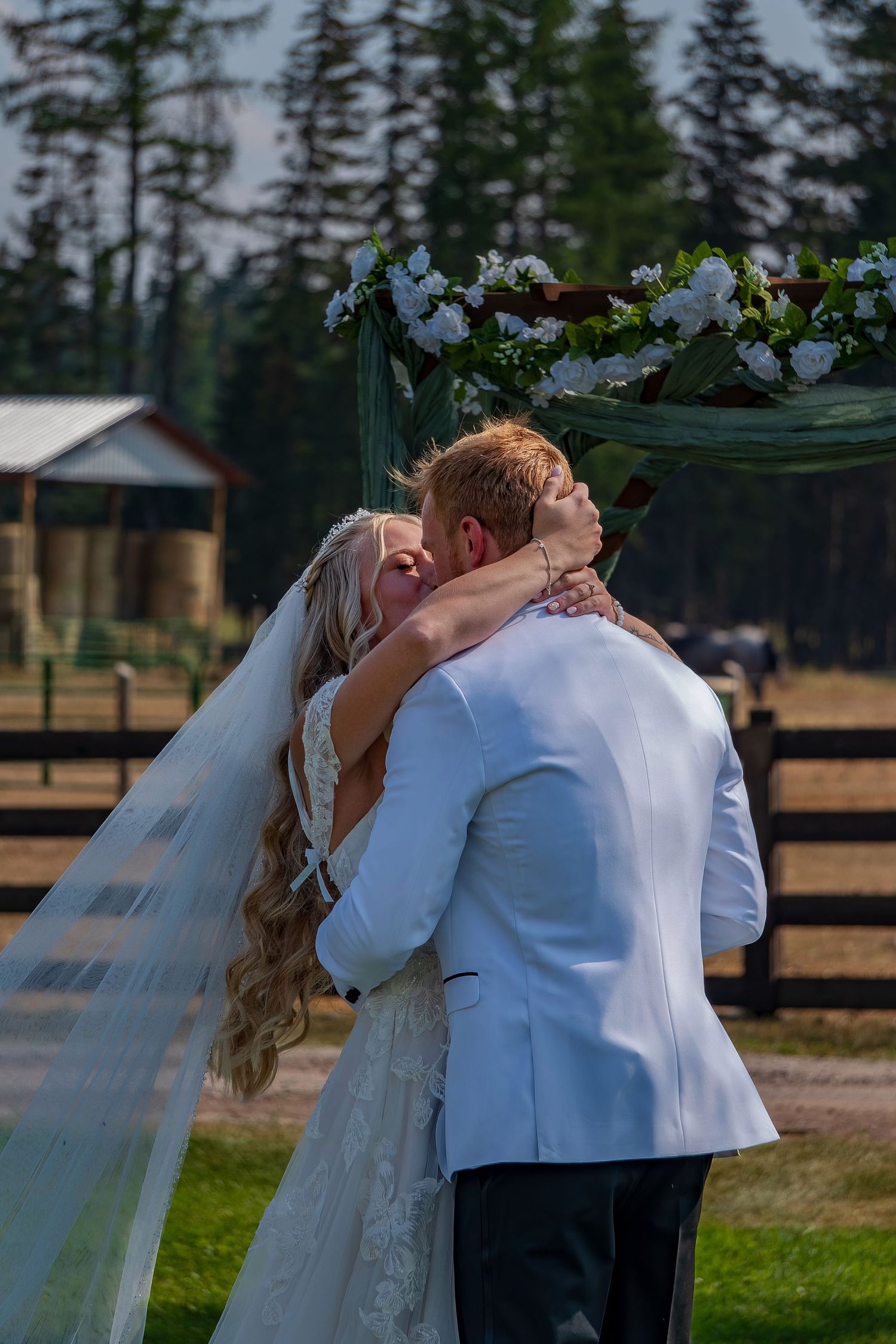 Romantic first kiss between newlyweds at rustic outdoor wedding venue with wooden fence.