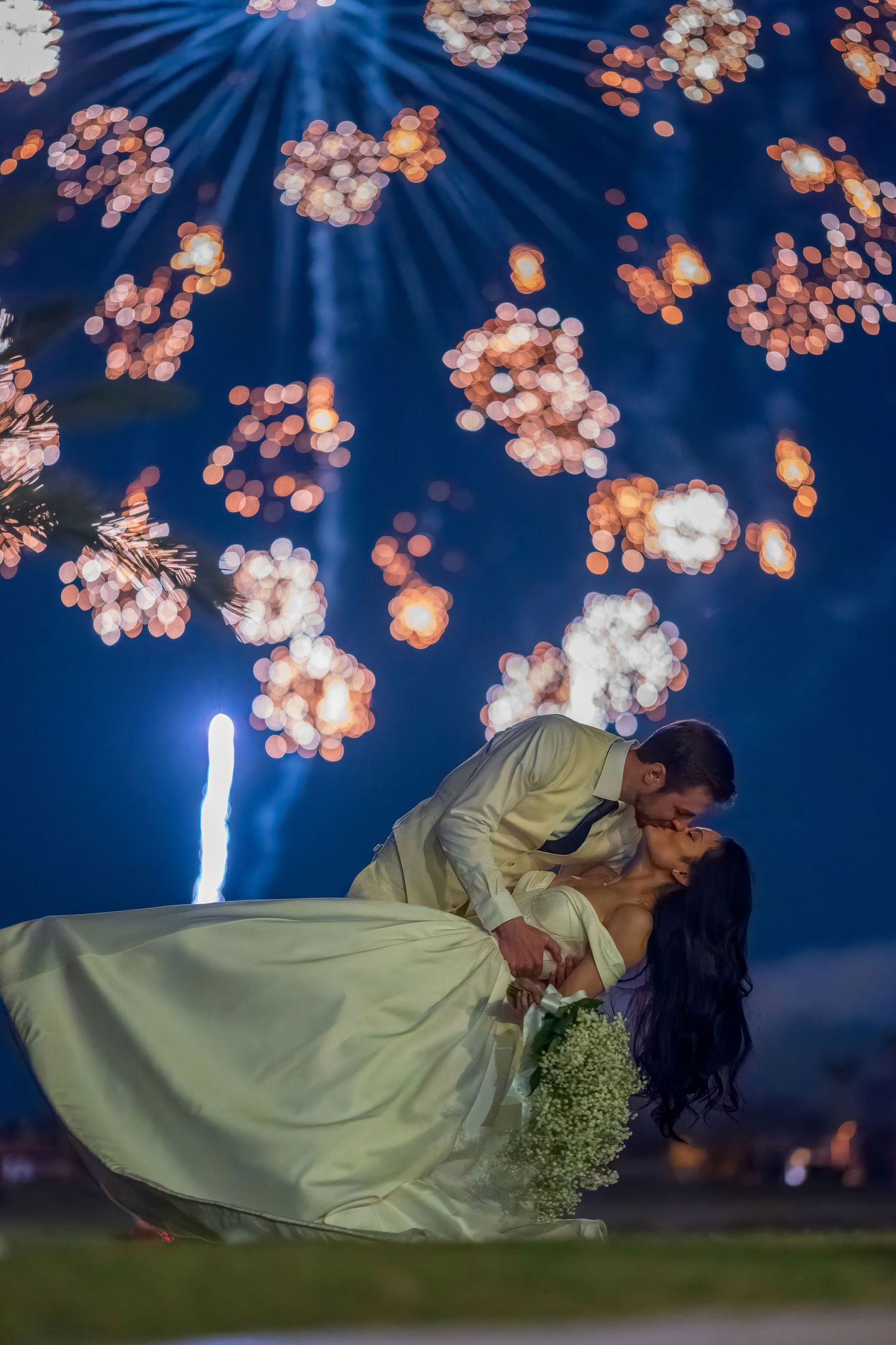Newlyweds pose for dramatic dip kiss photo with sparkling blue fireworks overhead.