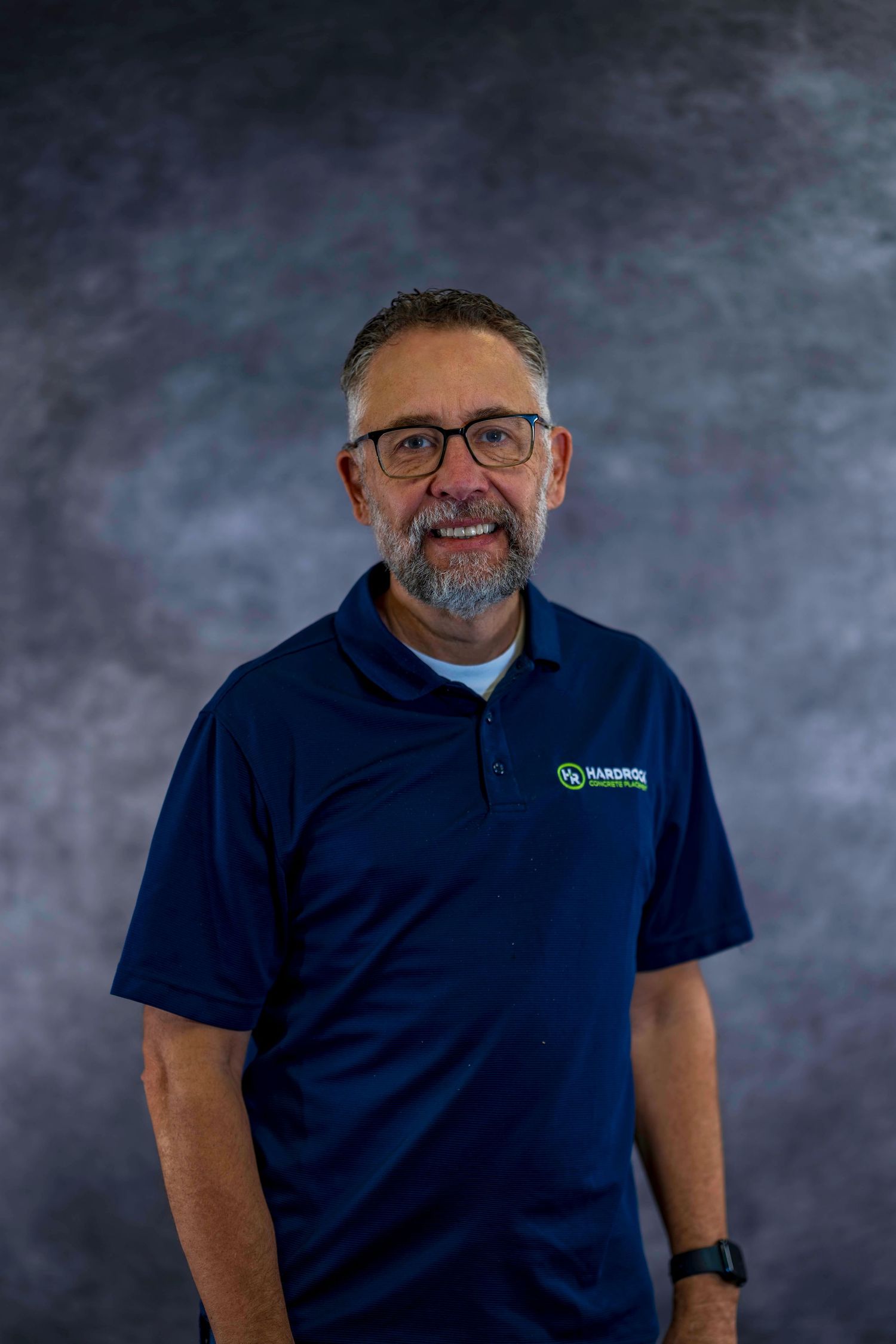 Professional portrait of person with beard wearing navy polo and glasses against gray background.