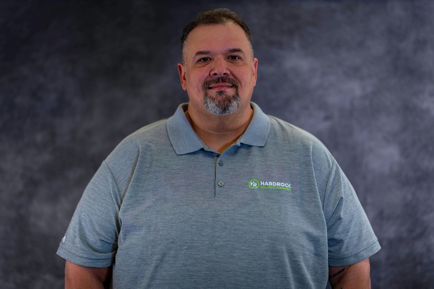 Employee portrait in gray polo shirt with embroidered logo against textured backdrop.