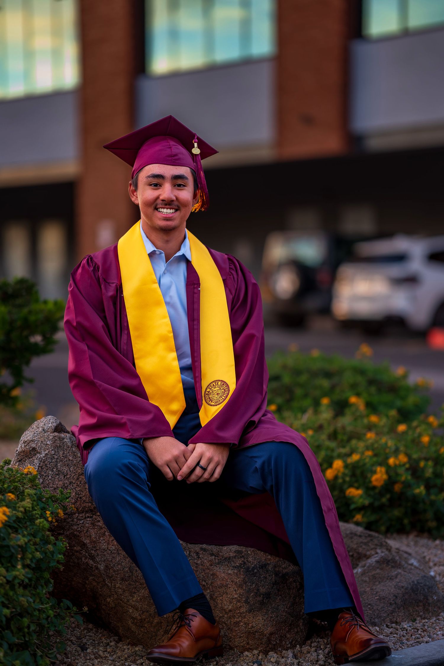 Graduate in maroon and gold stole sits on rock ledge near flowering bushes.