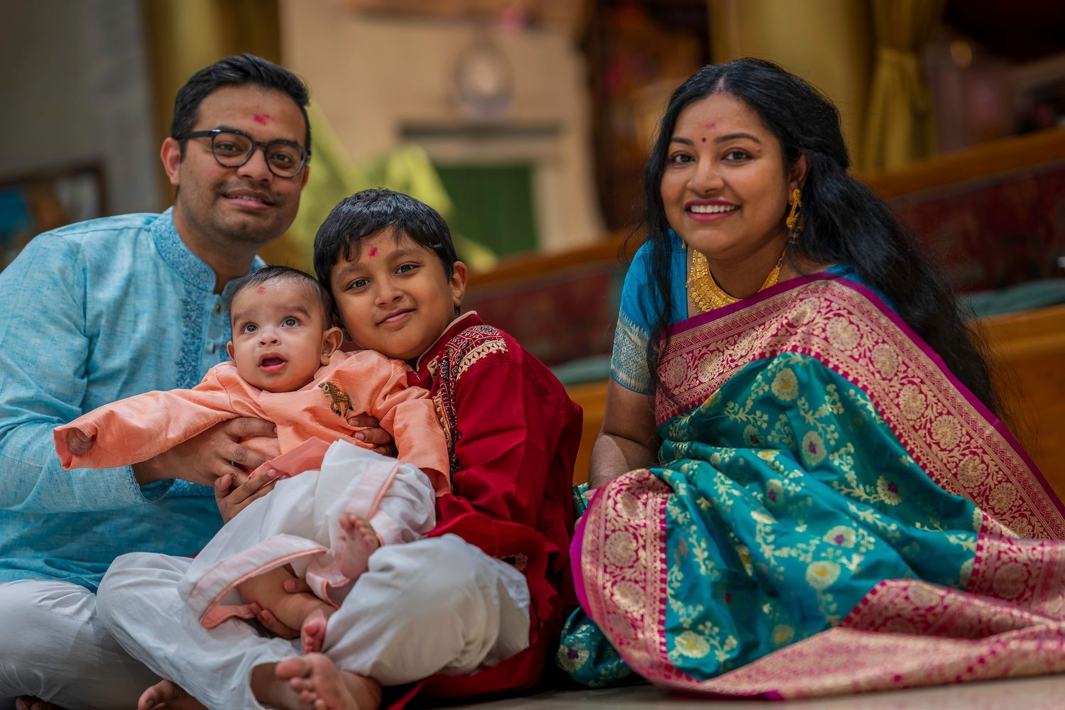 Indian family in traditional attire smiling together, parents with two young children in a festive home setting.