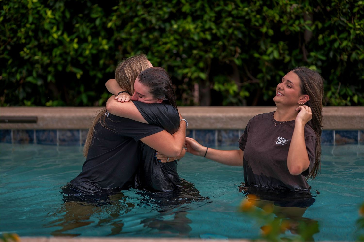Two women hugging in a pool after a baptism, with a third woman smiling beside them.
