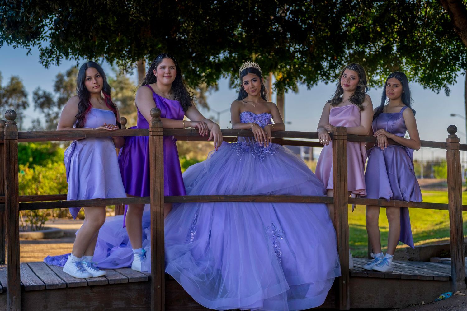 Quinceañera girl in lavender gown poses with four chambelanes on wooden bridge under tree.
