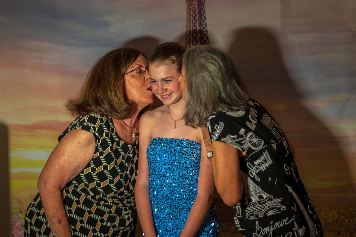 Two women kissing a young girl in a blue sequin dress at a Paris-themed event.