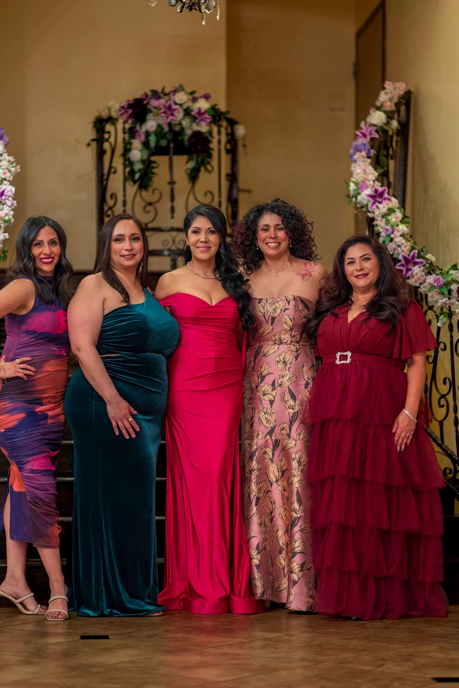 Five women in elegant formal gowns pose together smiling at a quinceañera celebration.