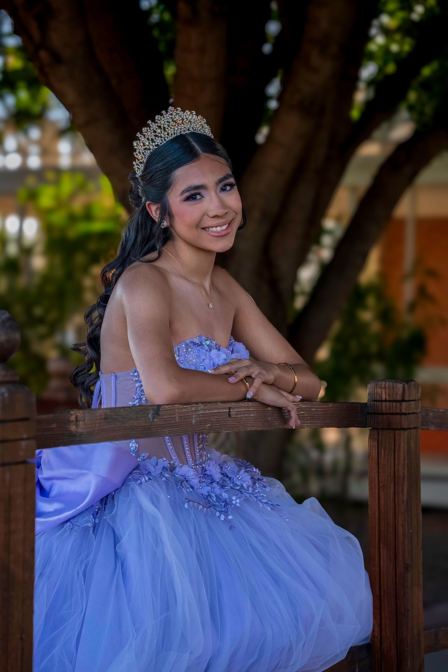 Smiling young Latina woman in purple quinceañera dress and gold tiara poses by wooden railing outdoors.