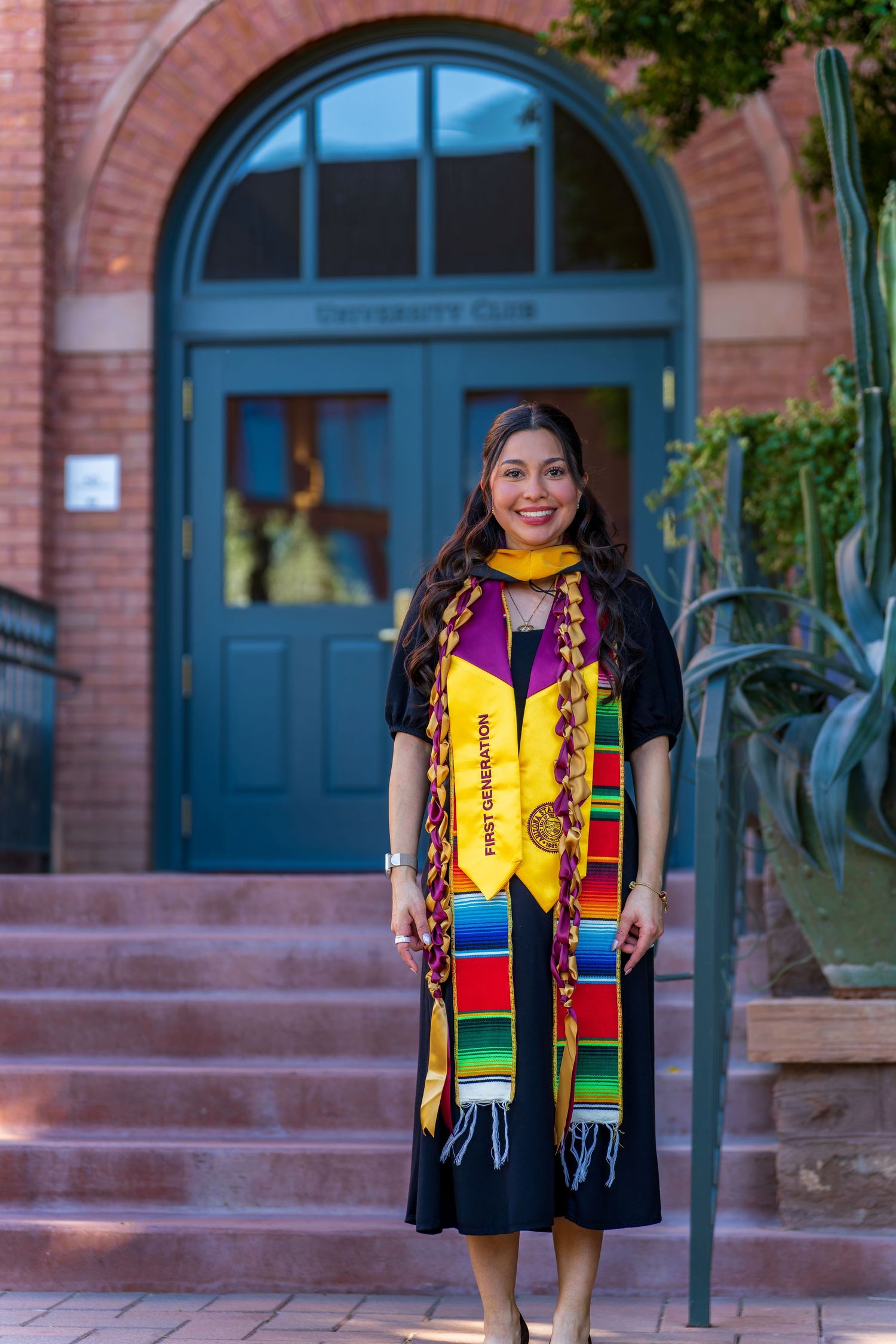 Graduate in black gown and colorful stole posing on brick steps with arched blue doors in background.