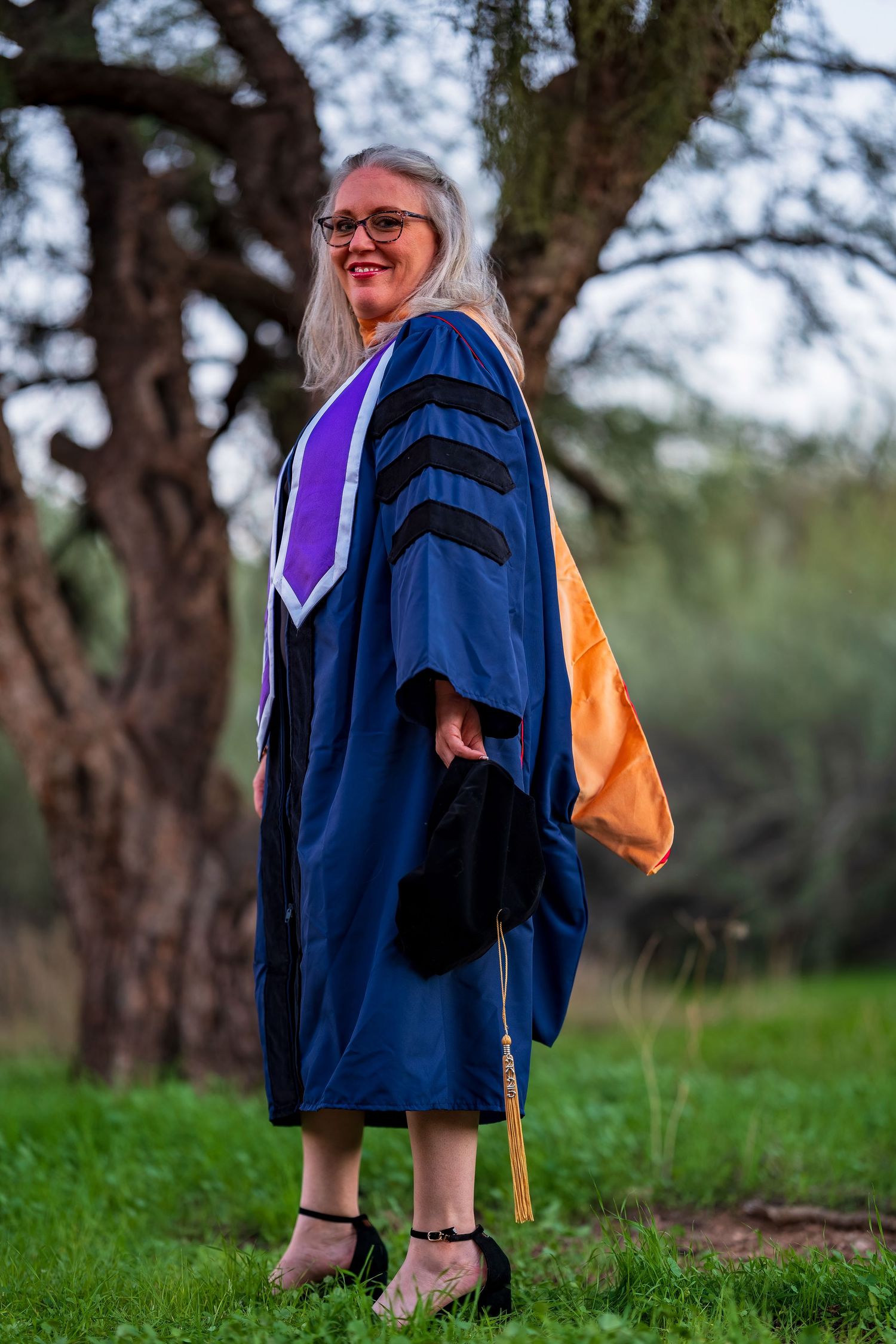 Graduate in blue doctoral regalia with purple stole standing on grass with trees in background.