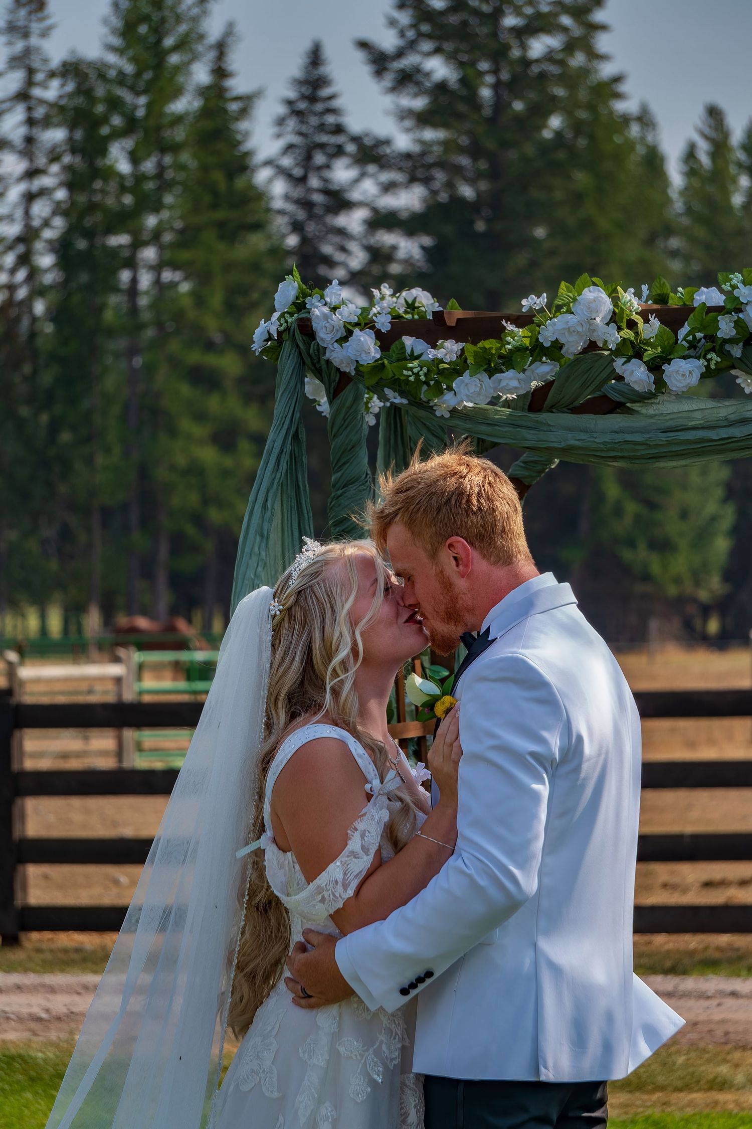 Intimate wedding kiss moment captured against backdrop of evergreen trees and rustic fencing.
