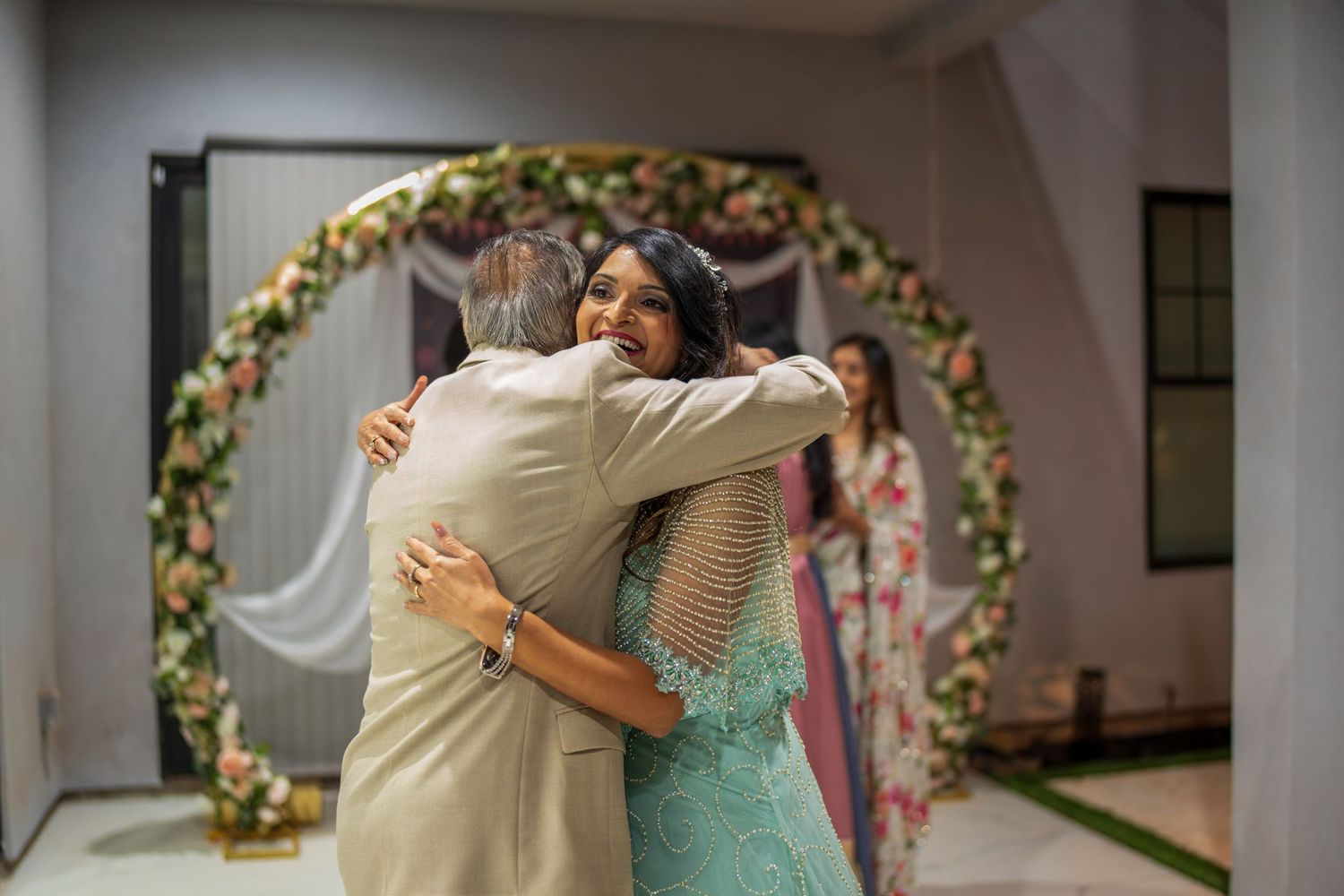 Guests embrace warmly at a celebration under an elegant floral arch with pink and white flowers.