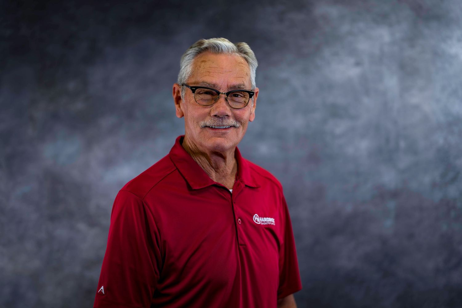 Professional portrait of person in red polo shirt and glasses against textured backdrop.