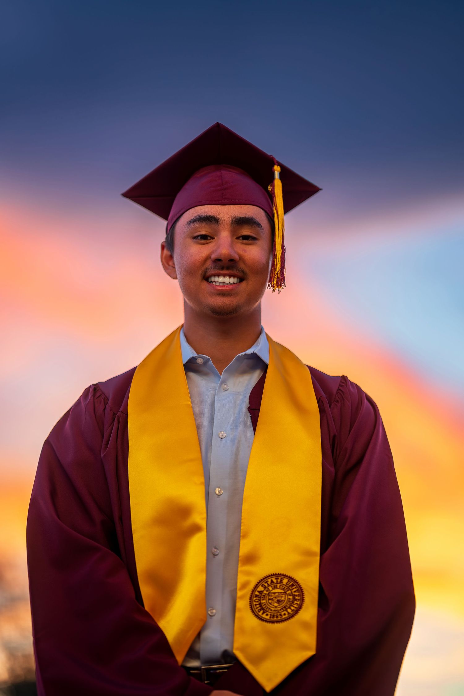 Graduate in maroon cap and gold stole smiles against colorful dusk sky.