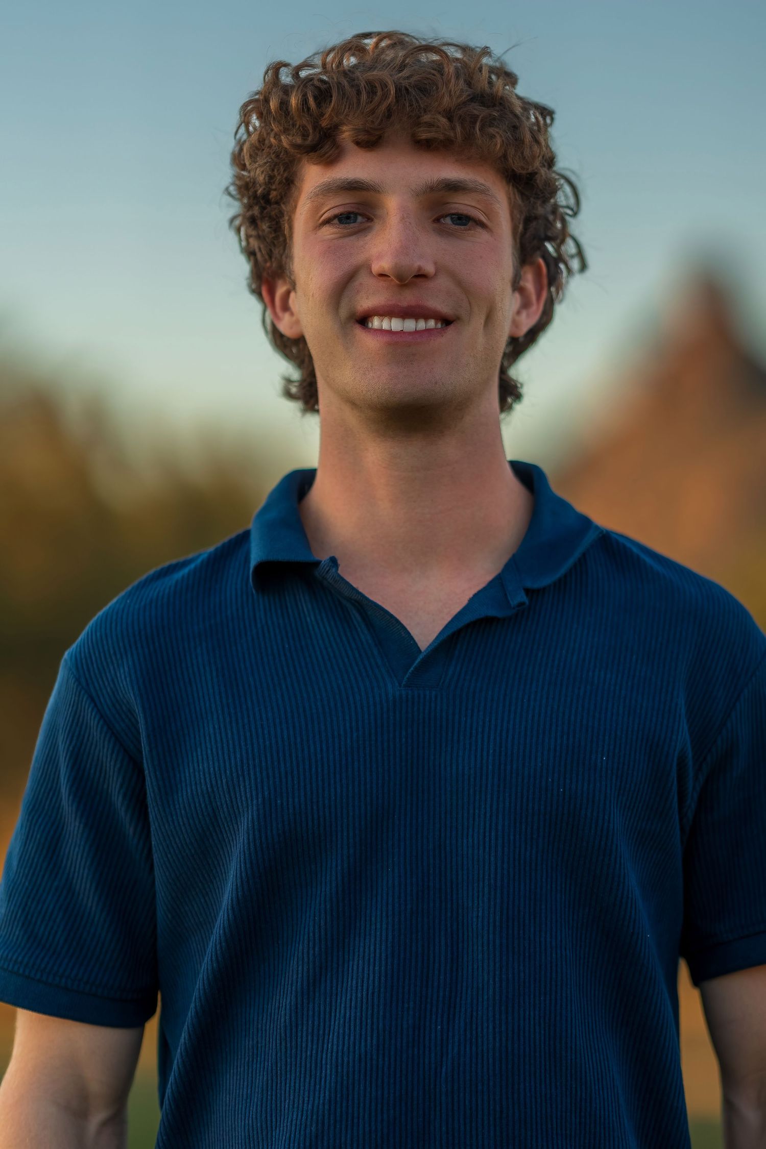Portrait of person in navy blue polo shirt with curly hair against evening sky.