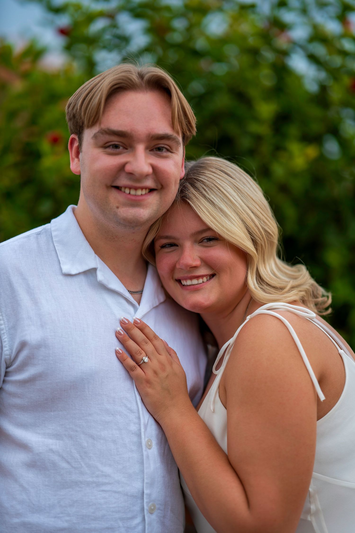 Happy couple smiling outdoors, woman in white dress with engagement ring, man in light blue shirt, green foliage background.
