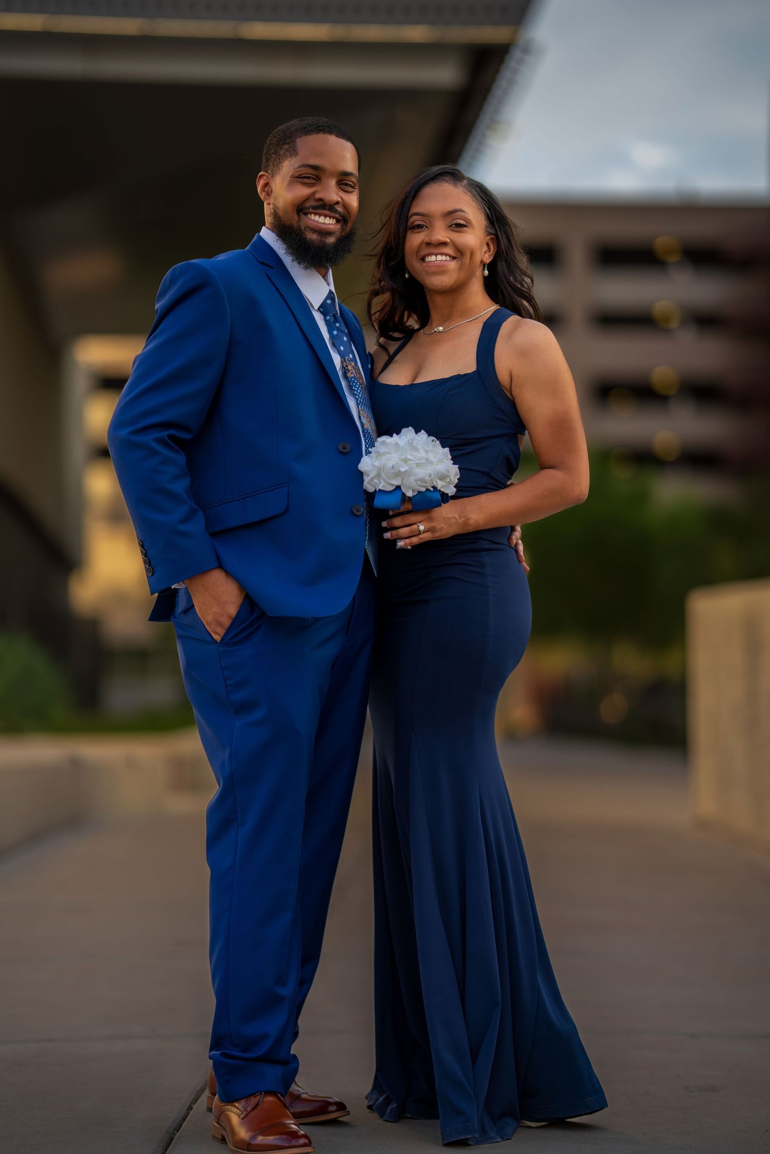 Smiling couple posing in coordinated royal blue suit and navy gown holding white floral bouquet outdoors.