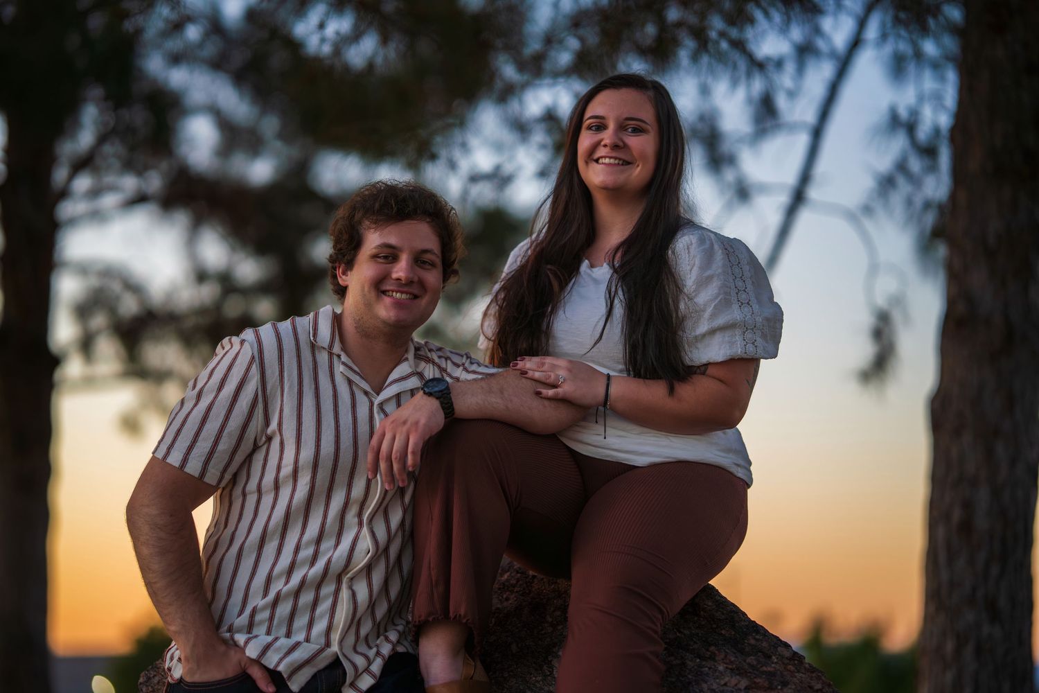 A smiling couple sitting together on a rock at sunset among pine trees in a warm evening glow.