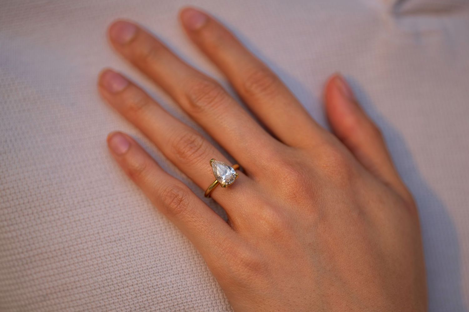 Close-up of a diamond solitaire engagement ring on a woman's hand resting on white fabric.