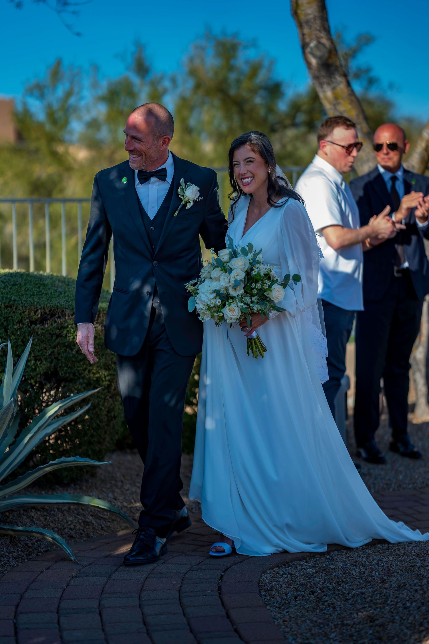 Bride in white gown holding bouquet walks with groom in navy tuxedo on brick path at outdoor wedding ceremony.