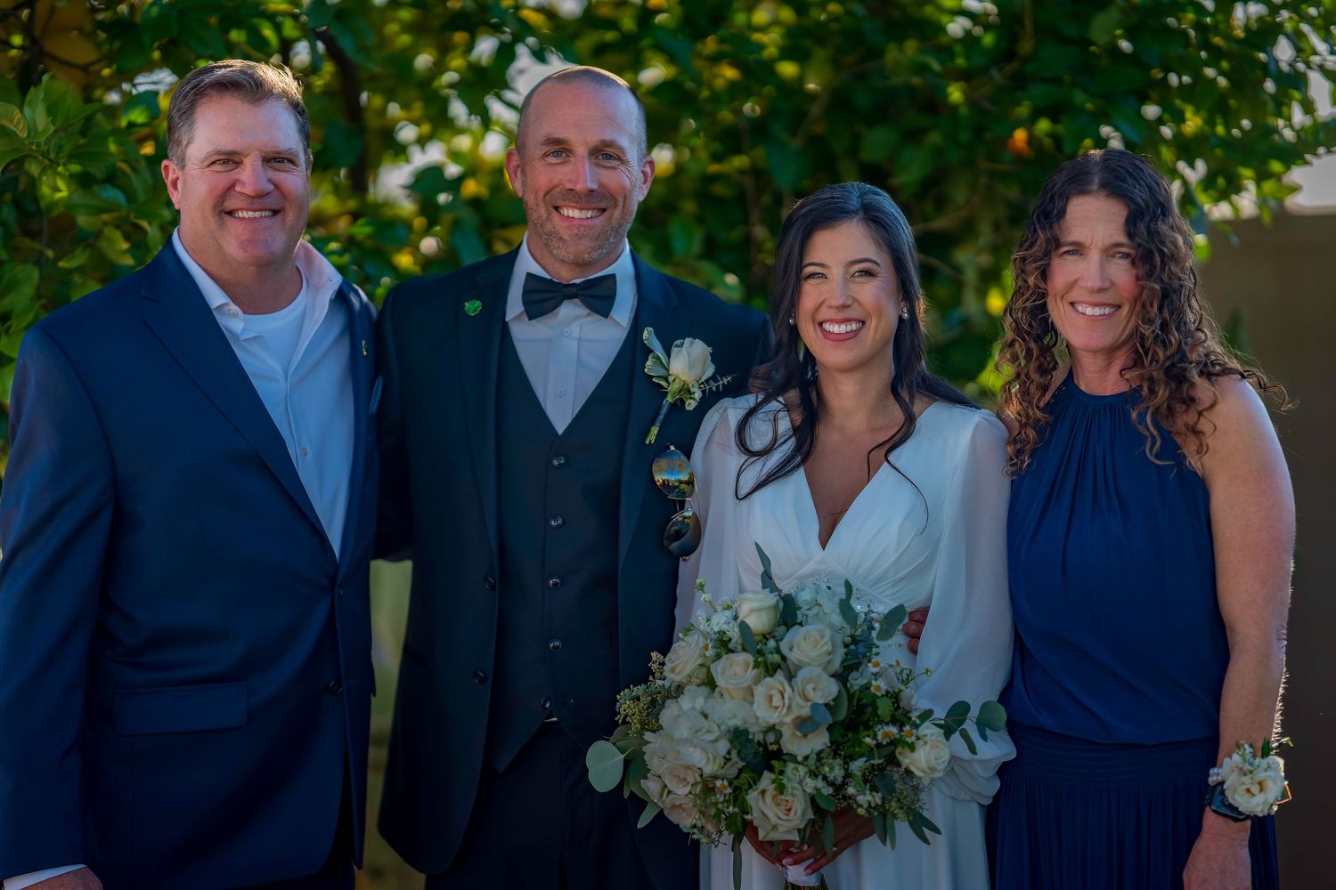 Bride and groom smiling with two guests in navy attire at outdoor wedding celebration.
