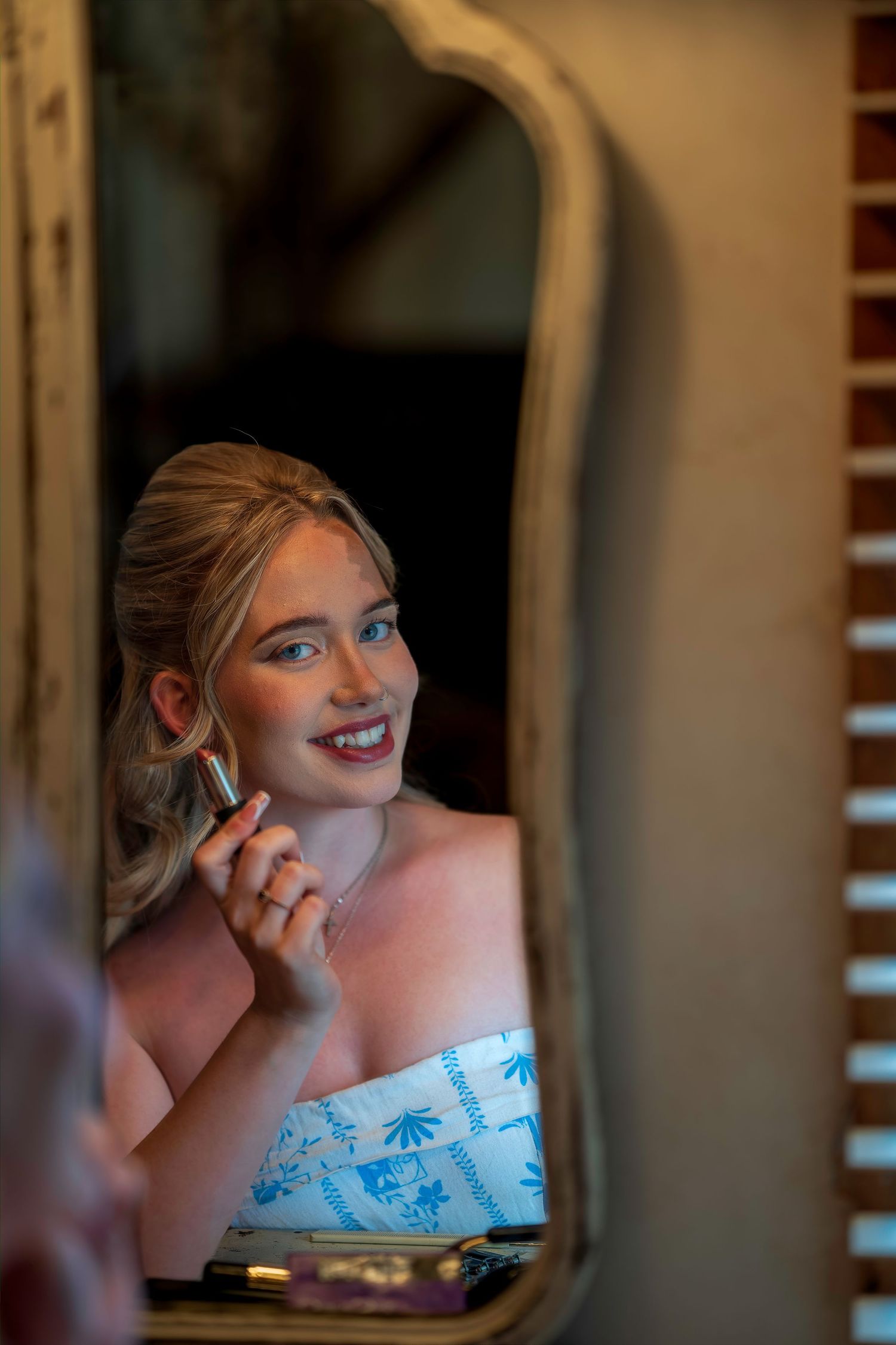 Smiling bride applying lipstick while getting ready, reflected in an ornate vintage mirror.