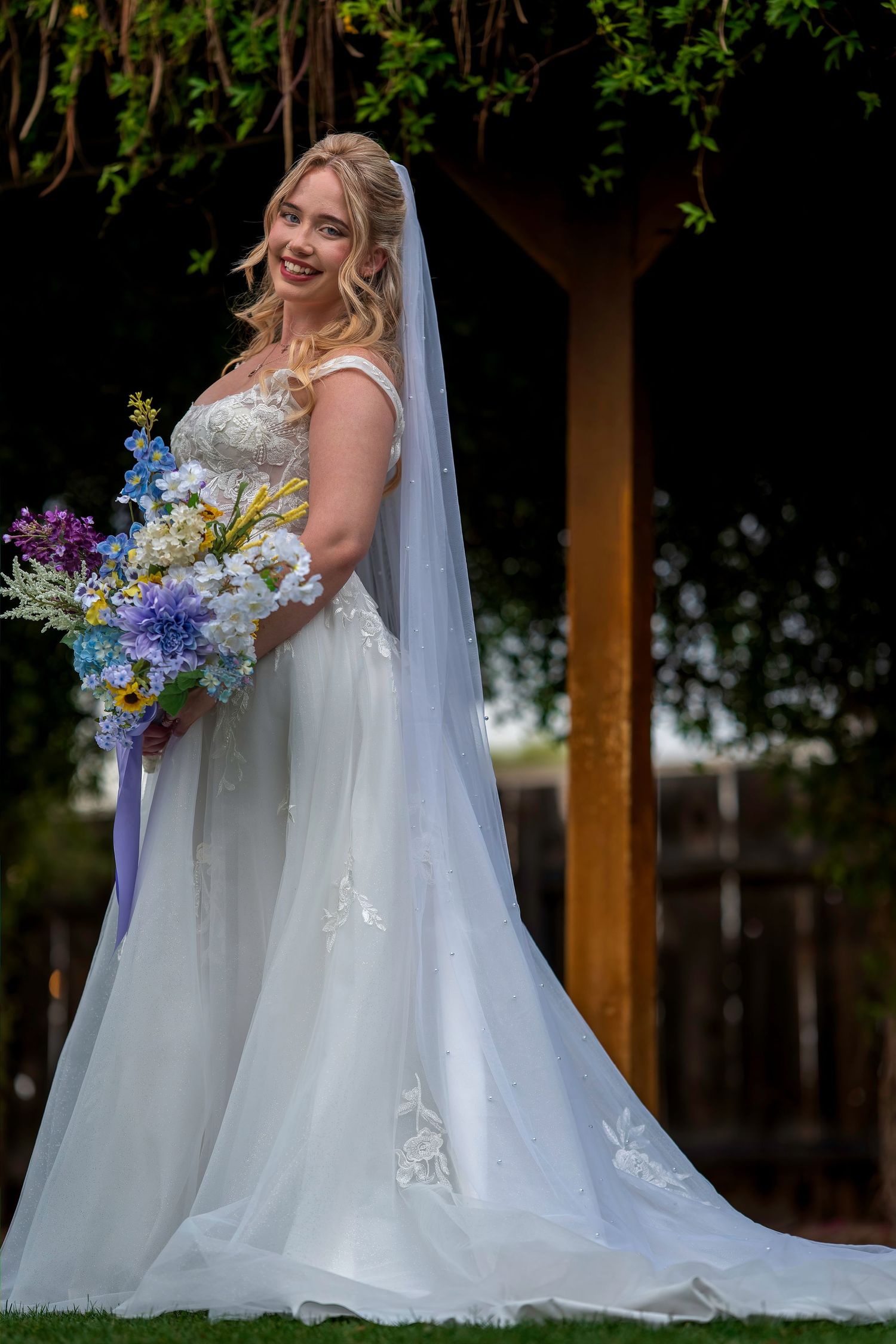 A radiant bride in a white lace gown with a long veil holding a colorful bouquet under a garden arbor.