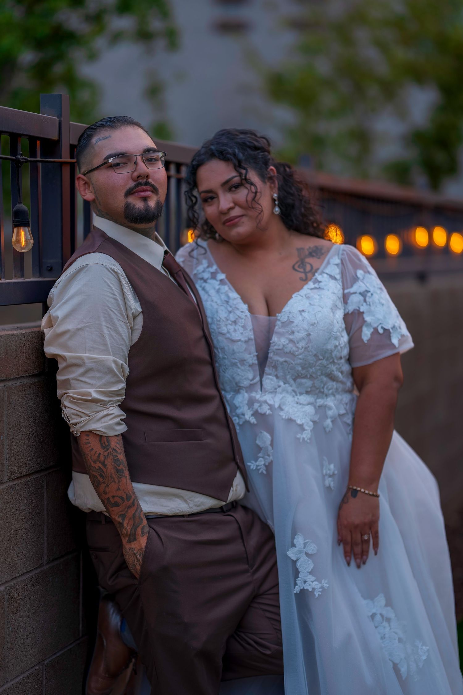 Bride in lace wedding gown and groom in brown vest with tattoos pose against a wall at dusk.