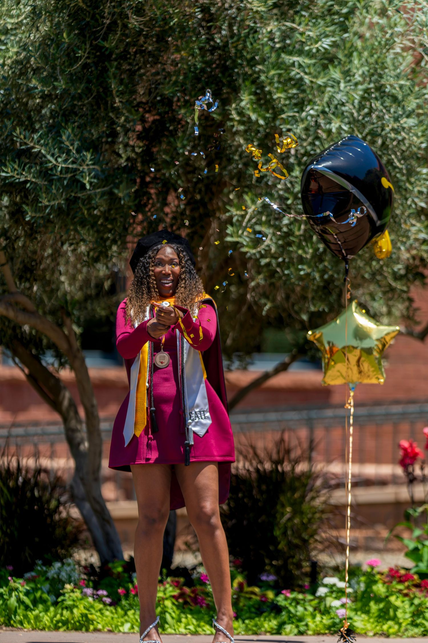 Happy graduate in maroon dress with stole poses outdoors holding graduation balloons in bright sunlight.
