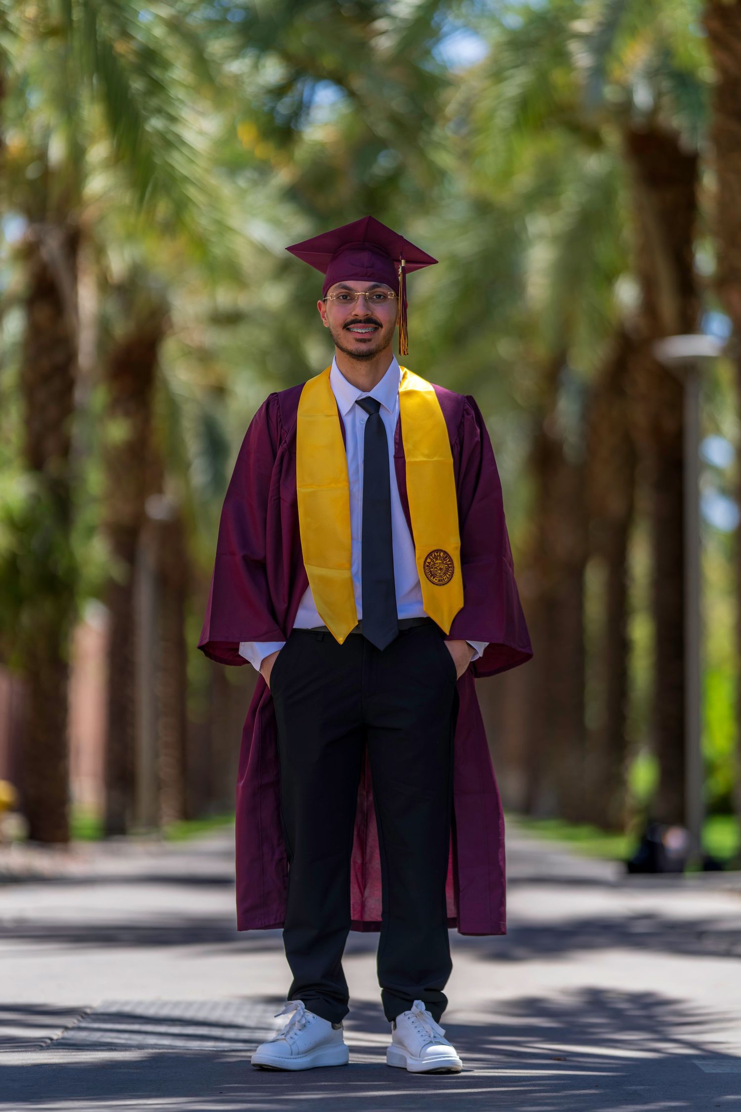 Smiling graduate in maroon cap and gown with gold stole standing on palm tree-lined path.