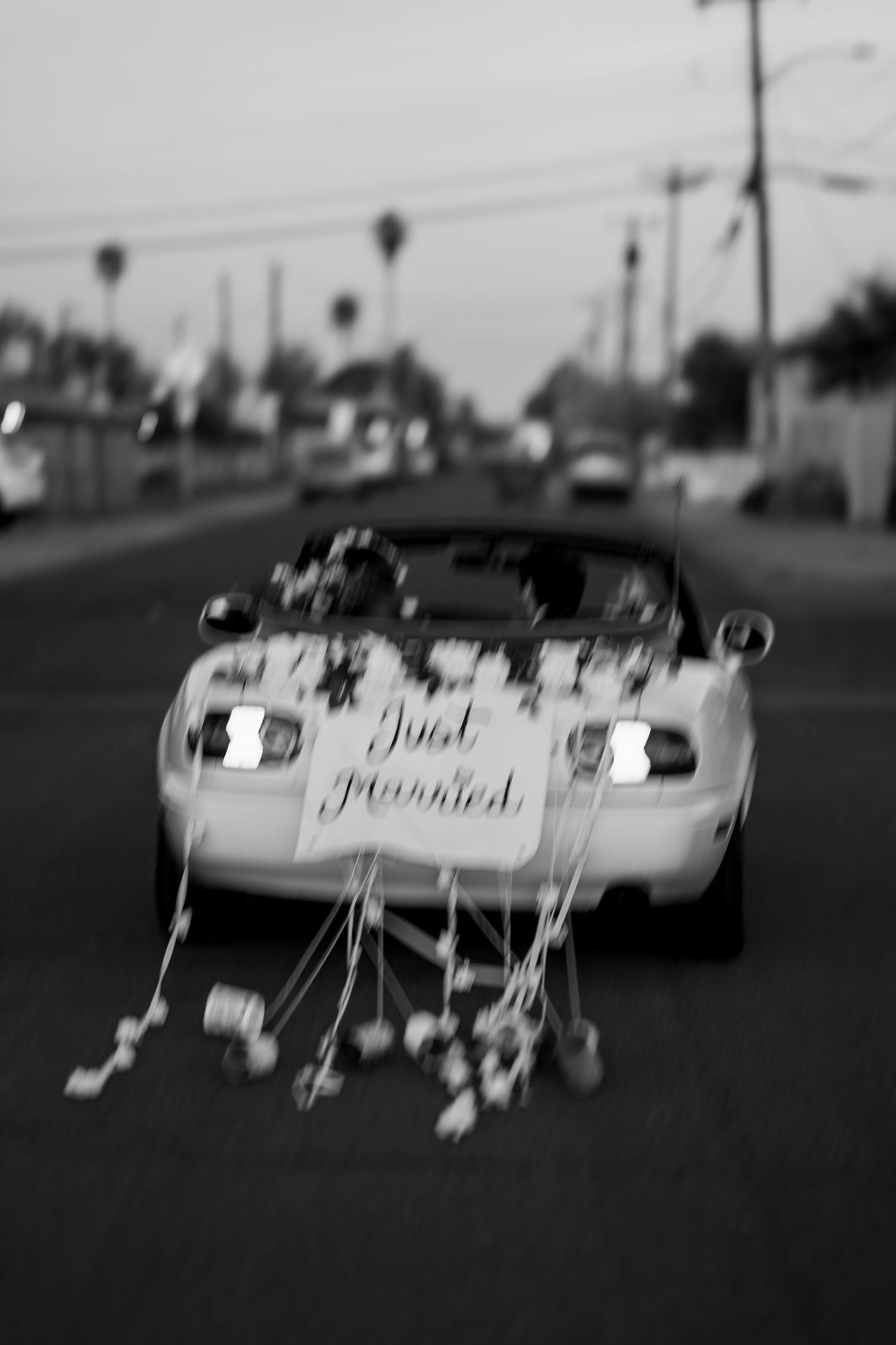 Black and white photo of a Just Married car decorated with flowers and ribbons driving down a street.