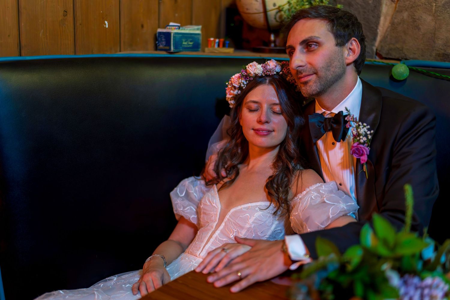 A bride in a floral crown rests on a groom in a suit, sitting together in a cozy booth on their wedding day.
