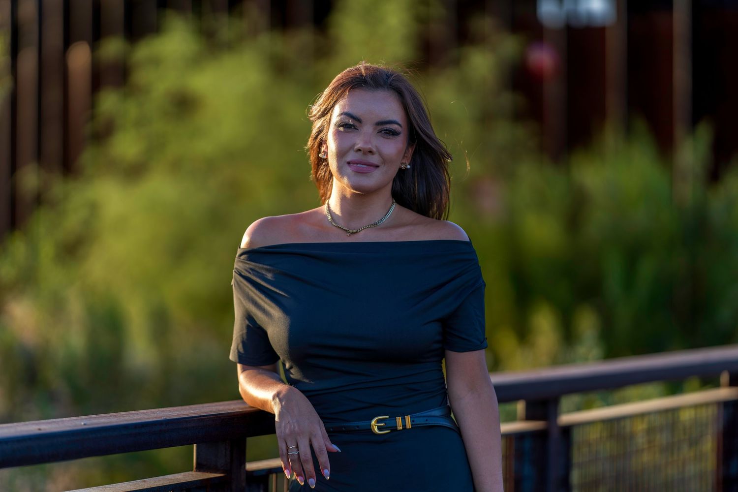 Woman in black off-shoulder dress with belt poses by railing with lush green foliage backdrop.