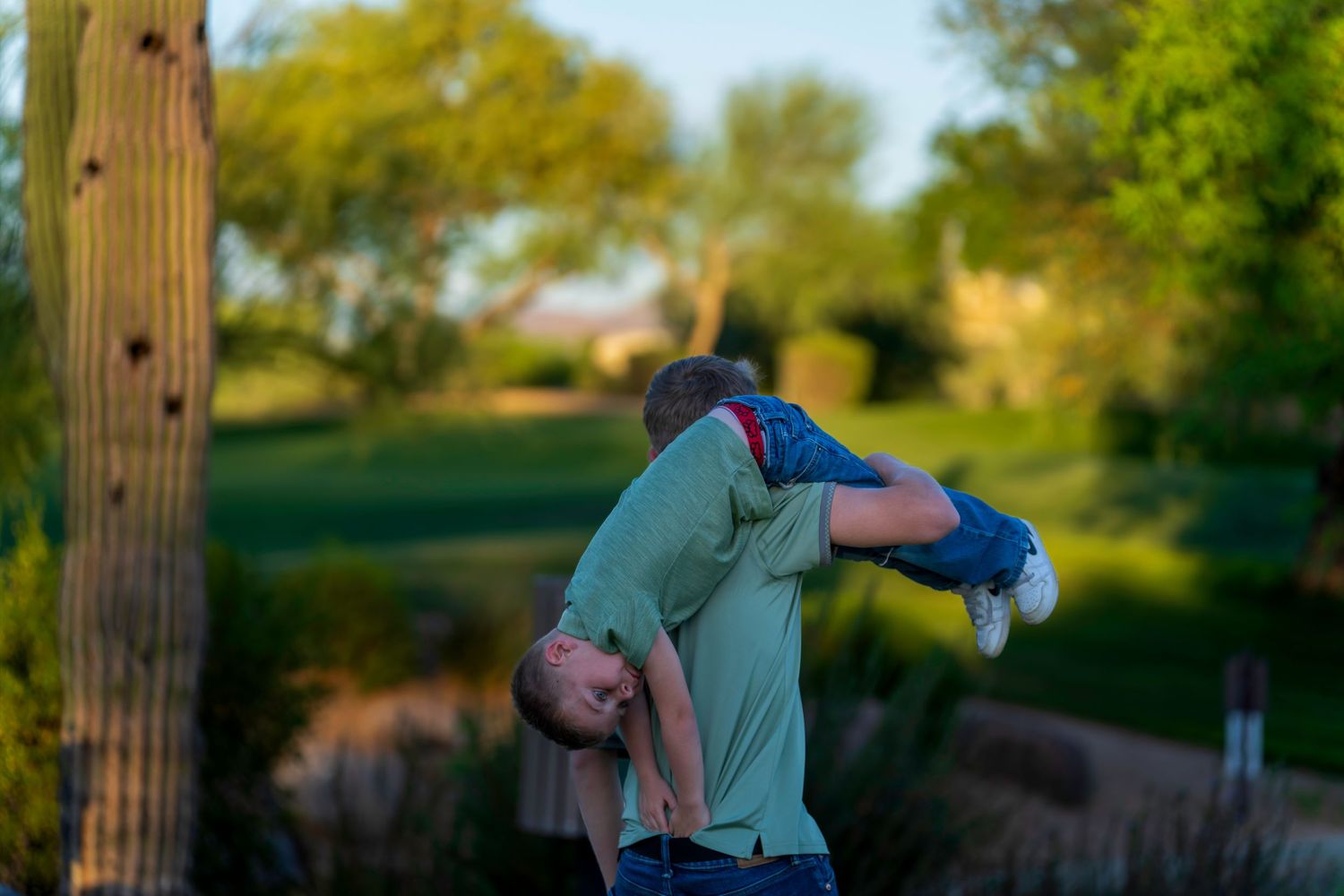 Father playfully lifting young child upside down outdoors near a saguaro cactus in warm evening light.