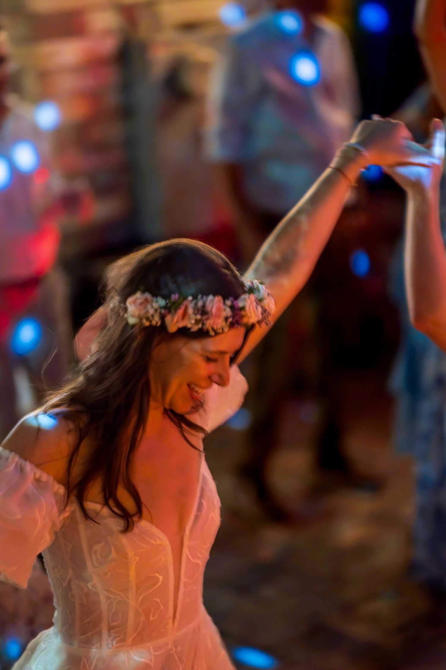 Smiling bride in white dress and floral crown dancing joyfully at a wedding reception with blue bokeh lights.