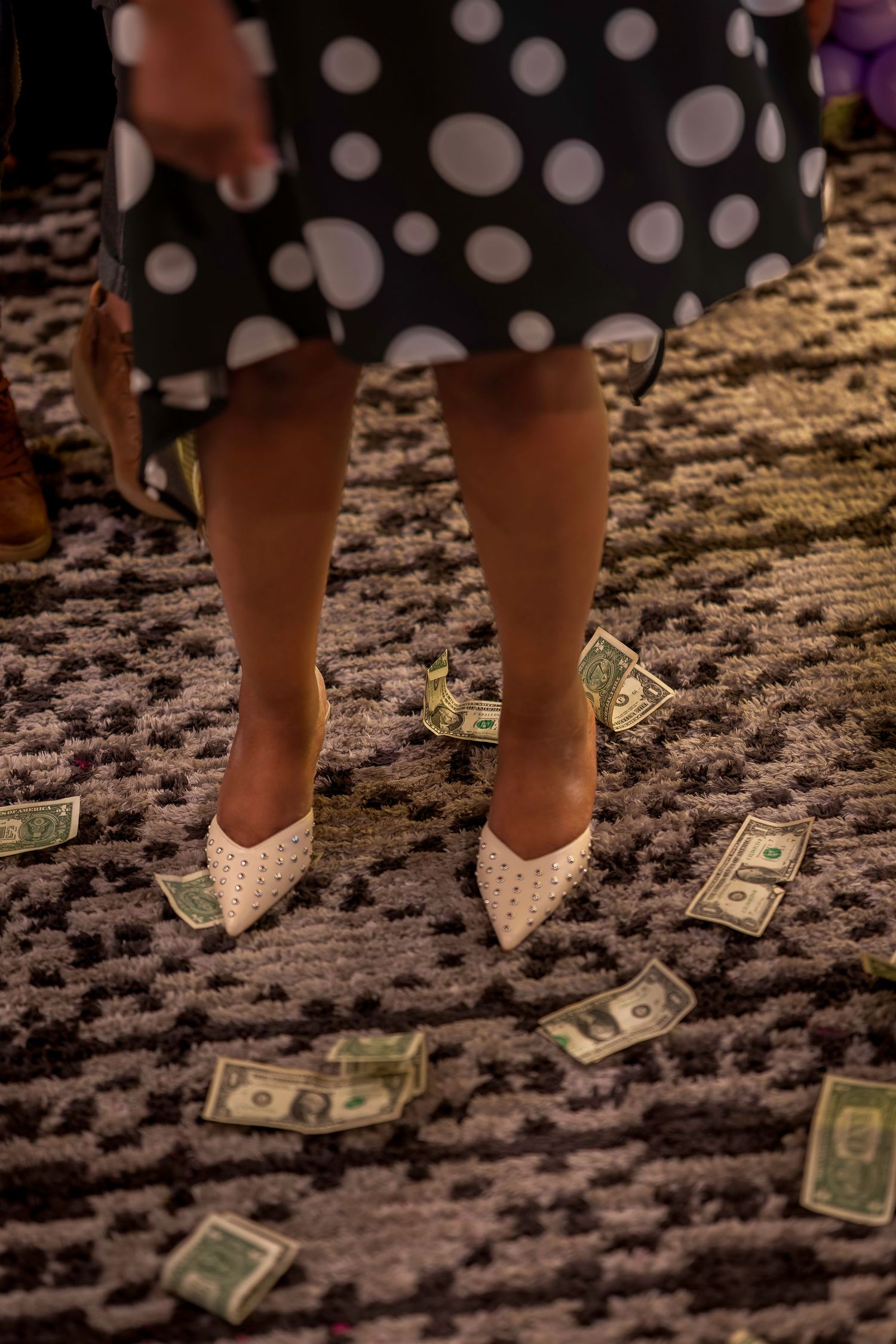 Woman in polka dot skirt and white heels standing on carpet surrounded by scattered US dollar bills.