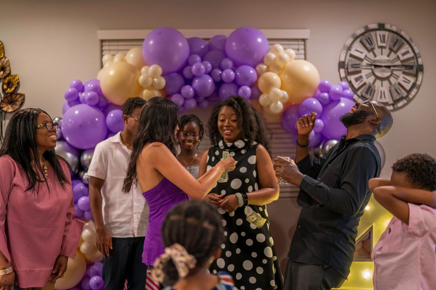 People dancing and celebrating at a party with purple and gold balloon arch decorations in a festive indoor setting.