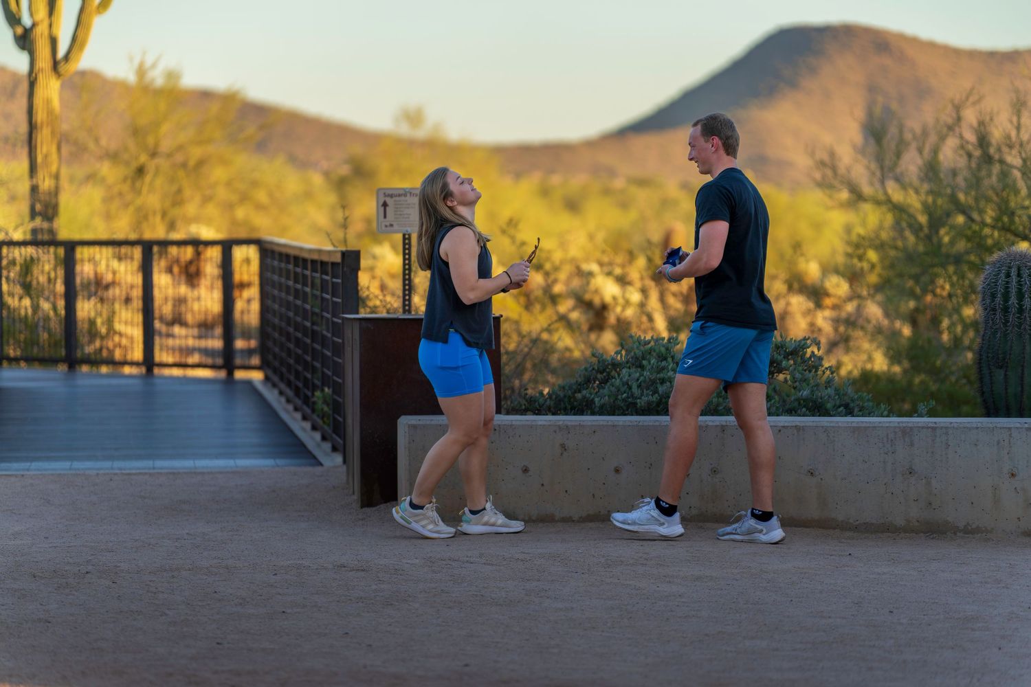 Two people in blue shorts and black shirts jogging on a desert path with mountain views at sunset.