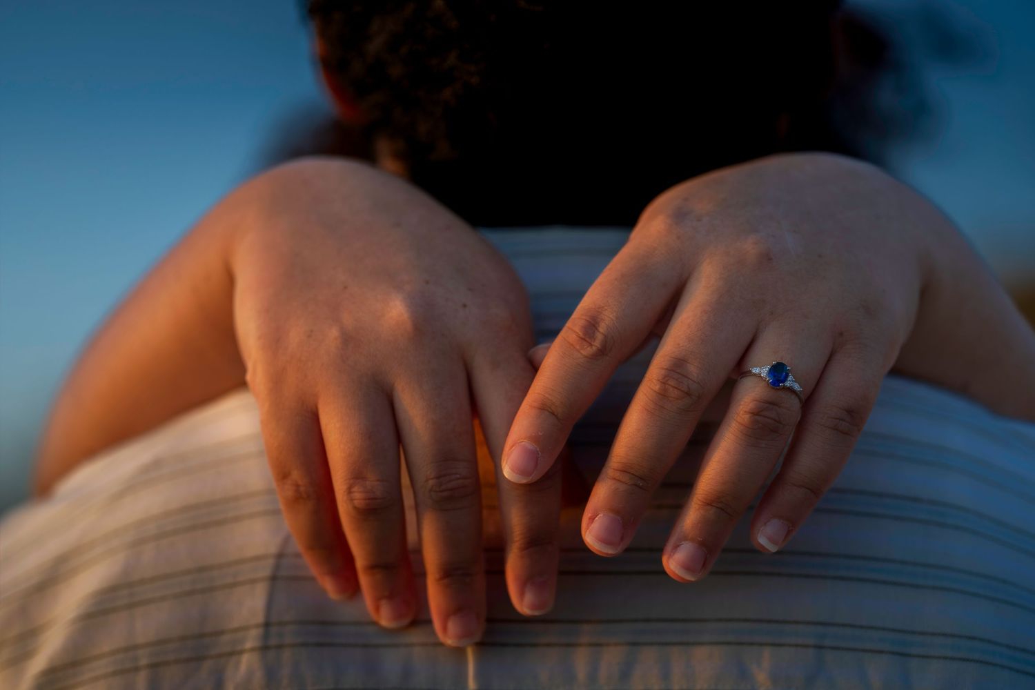 Close-up of hands showing sapphire and diamond engagement ring resting on mans striped shirt shoulder.
