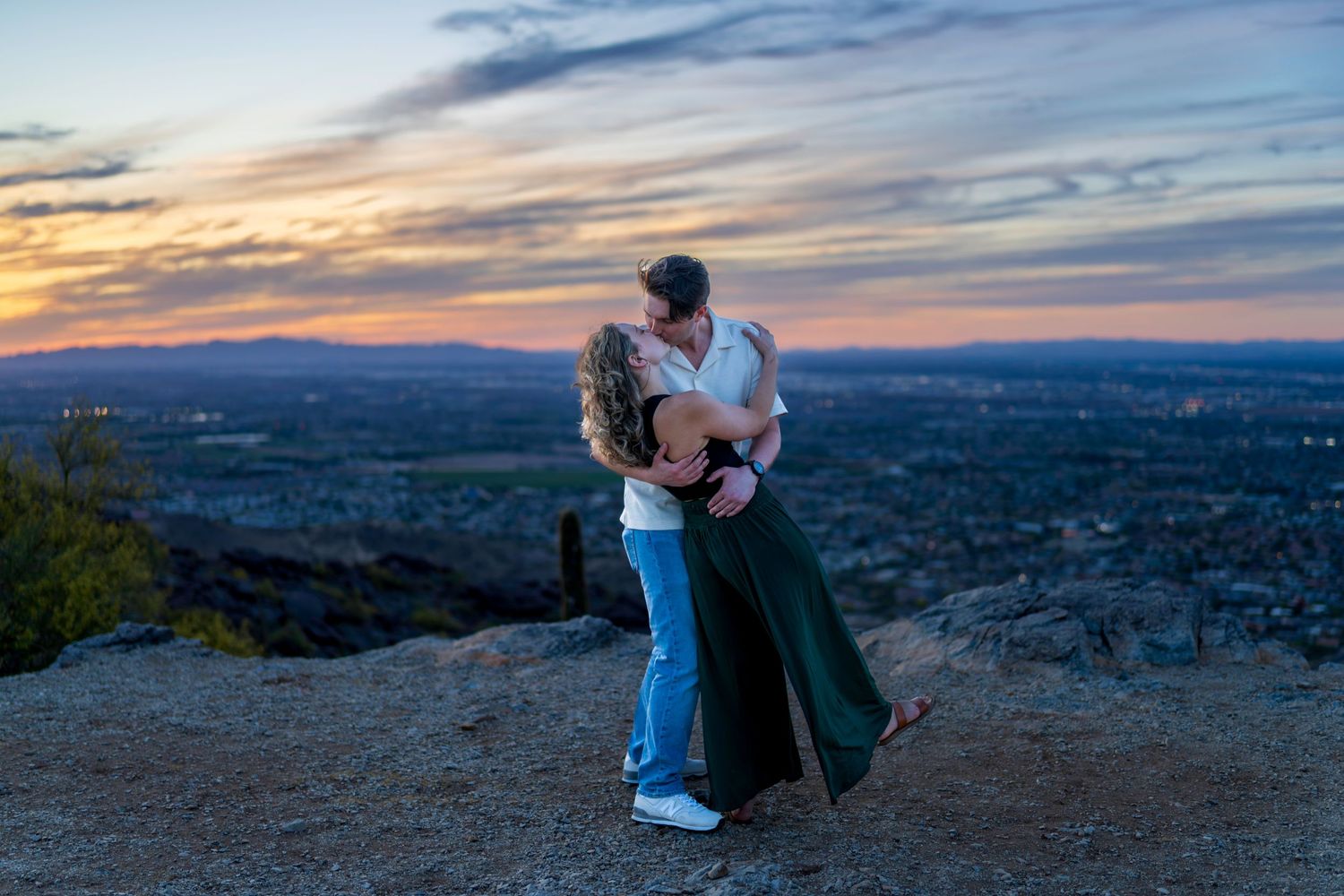 Couple kissing on rocky overlook at sunset with city lights visible in the background below.